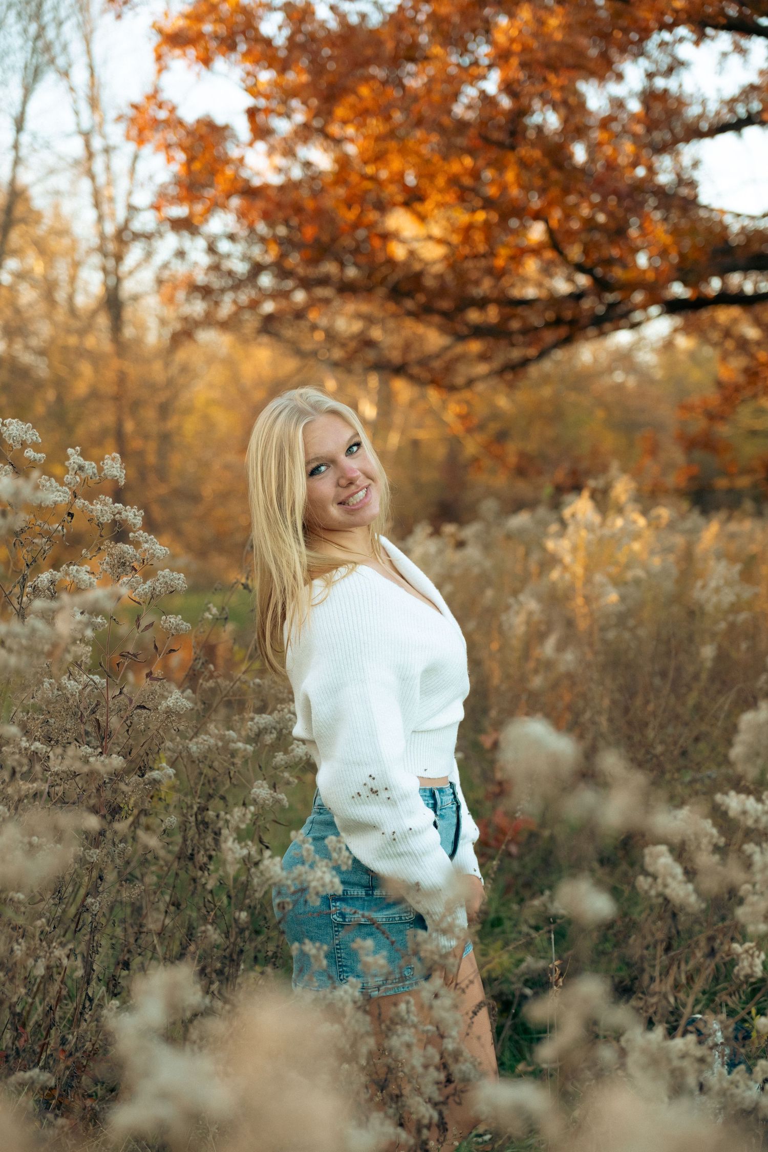 Smiling blonde woman in white sweater and denim skirt posing among wildflowers in autumn forest.