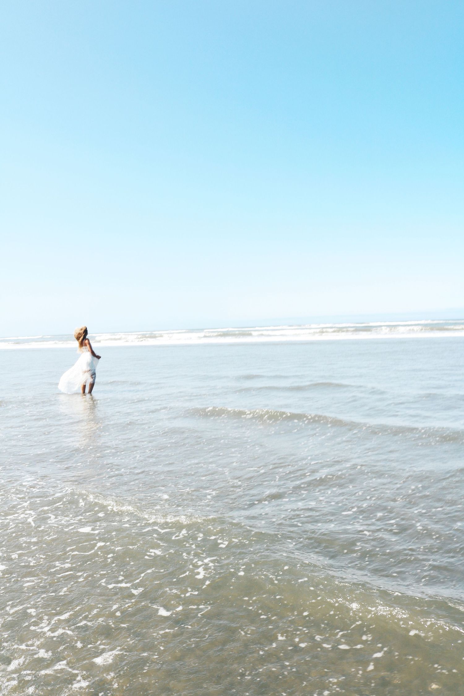 Woman in white dress and hat wading in ocean waves under clear blue sky.