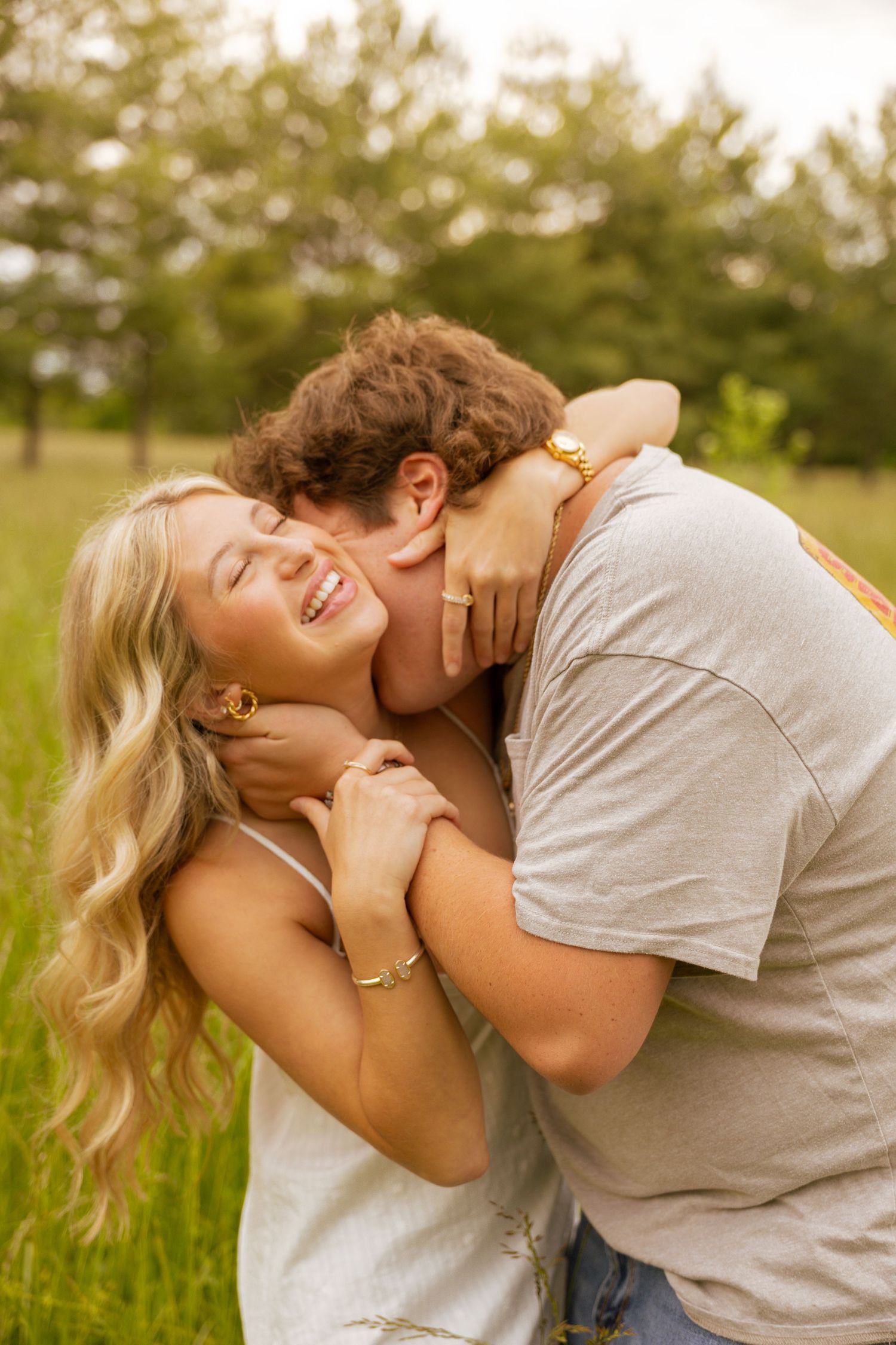 A young couple embraces outdoors, man kissing woman's cheek as she smiles joyfully in a lush green field.