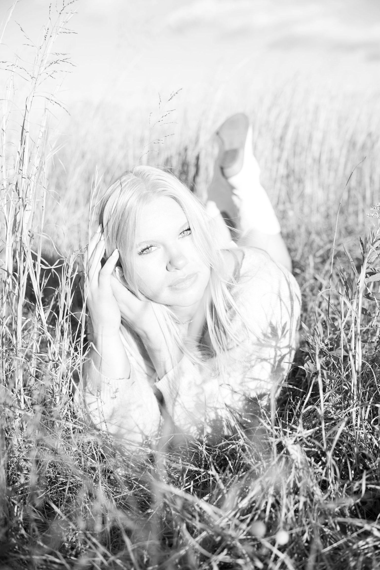Black and white photo of a smiling blonde woman lying in tall grass fields, looking up at the camera.