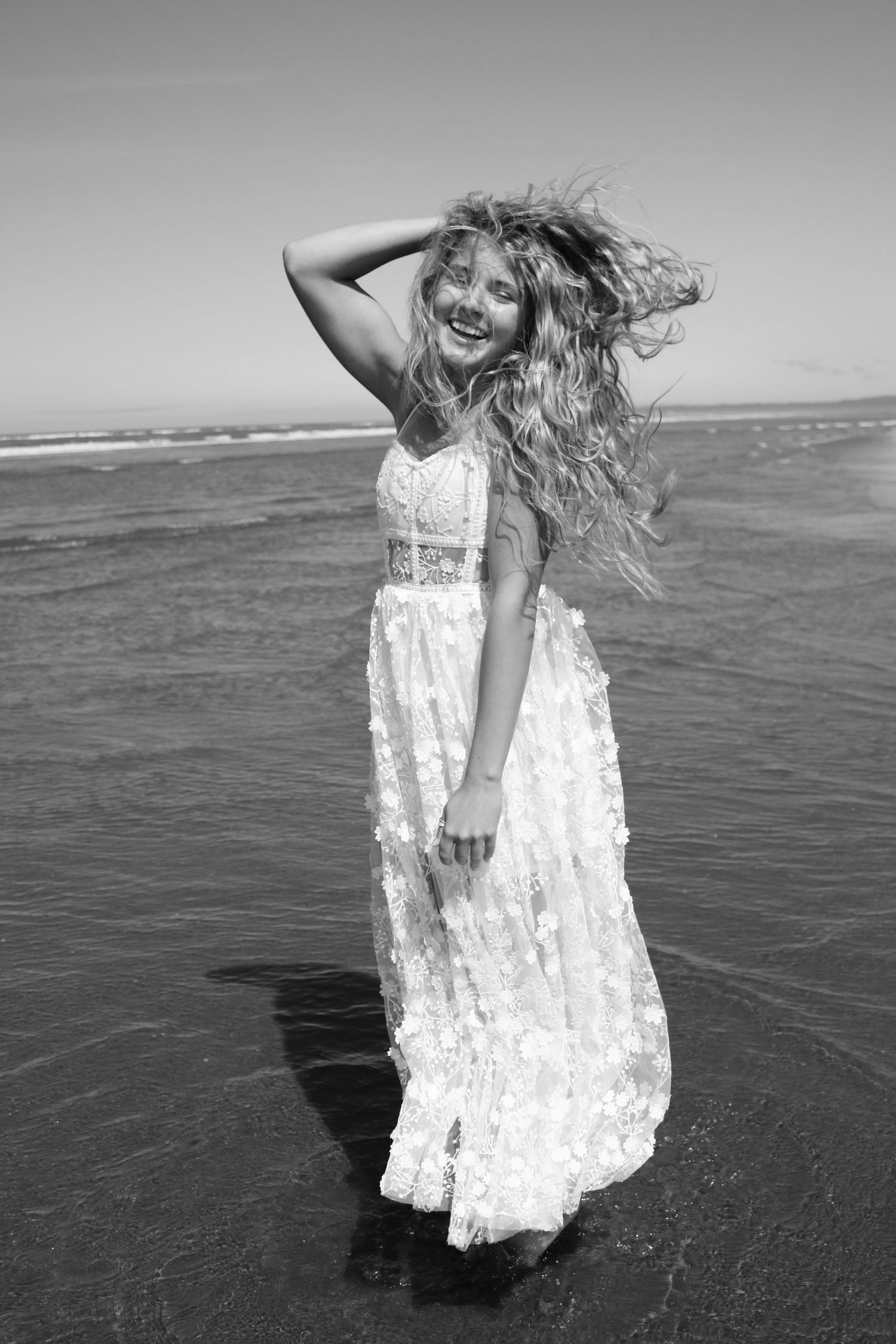 Black and white photo of a smiling woman in a white lace dress standing in ocean waves in Washington State with wind-blown hair.