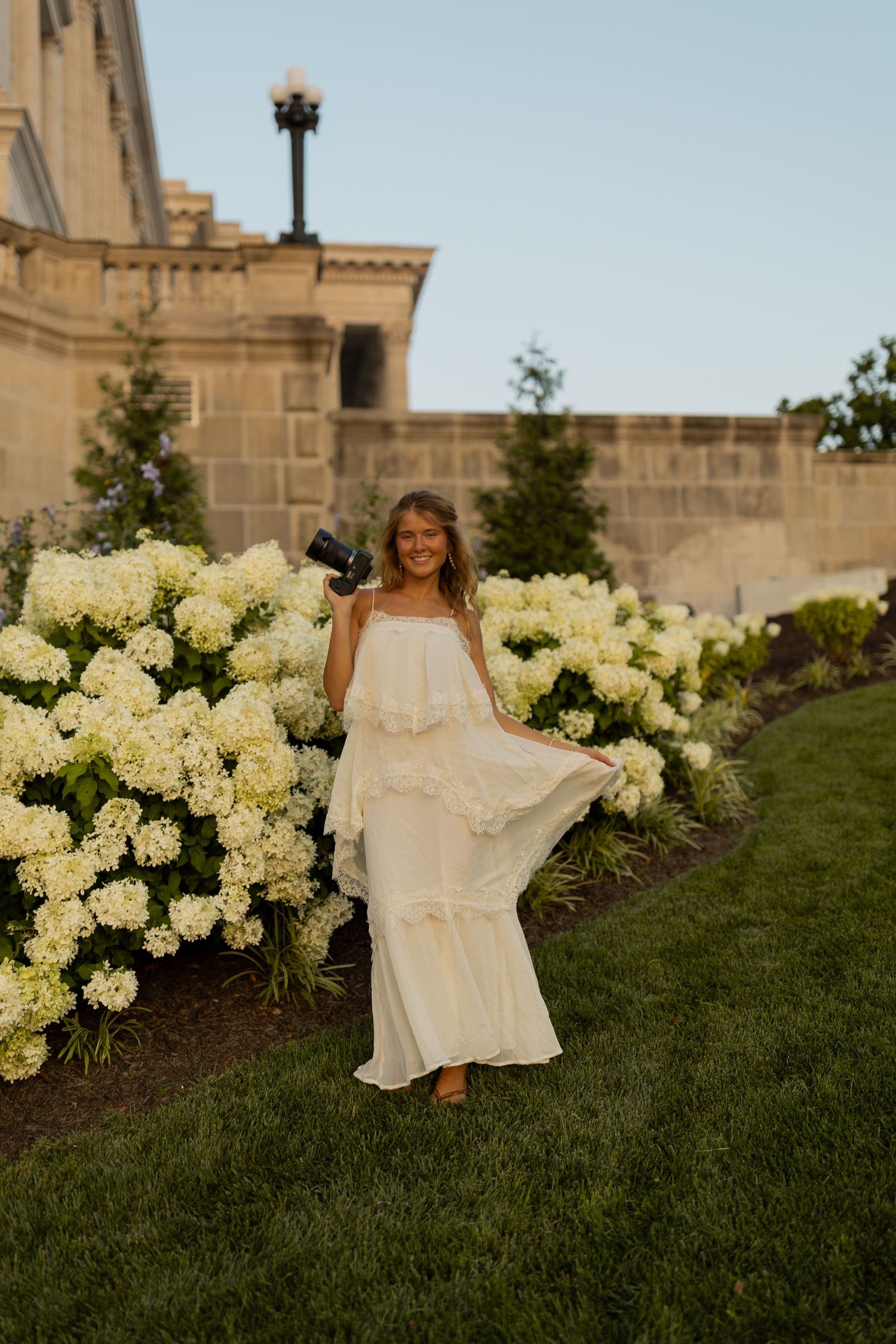 A smiling woman in a white maxi dress holds a camera near blooming white hydrangeas in a lush garden.