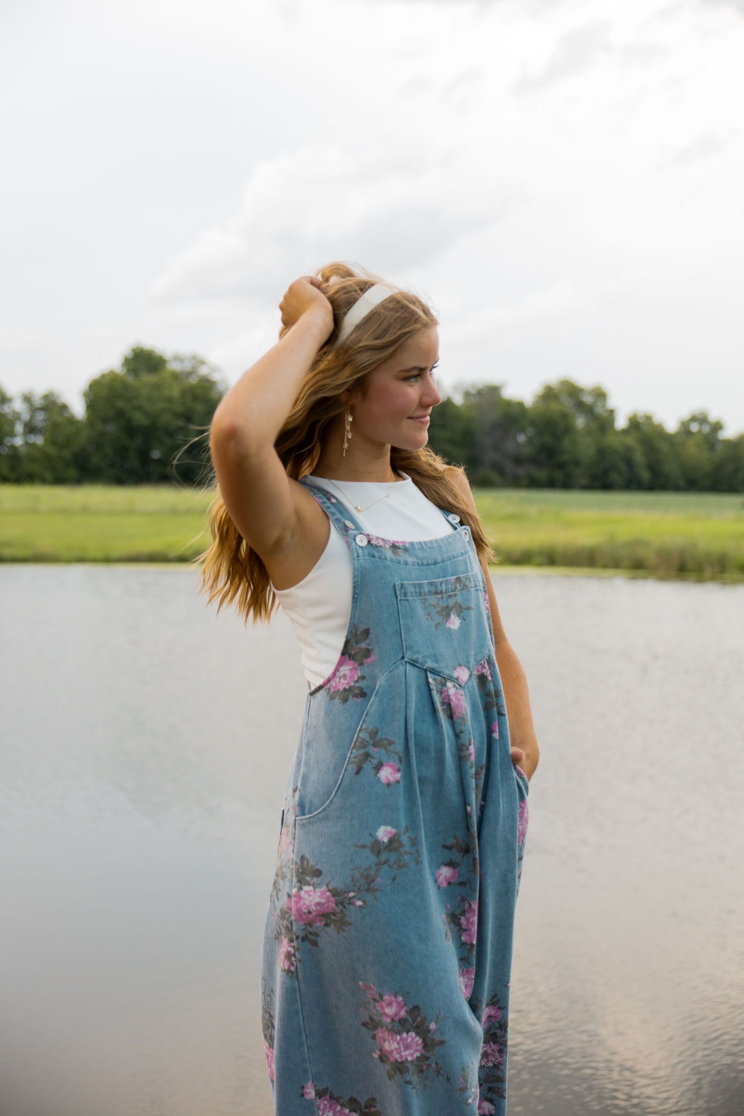 Young woman in floral overalls and white shirt smiles by a serene lake, touching her hair on a cloudy day.