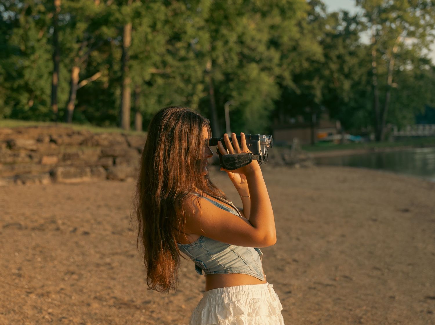 Young woman with long hair photographing with a camera on a sandy beach at golden hour.
