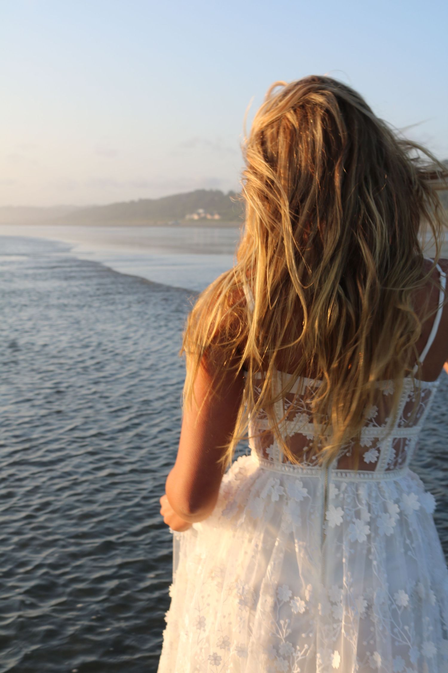 Young woman in a white floral lace dress standing on a beach at sunset with long blonde windswept hair in Washington State.