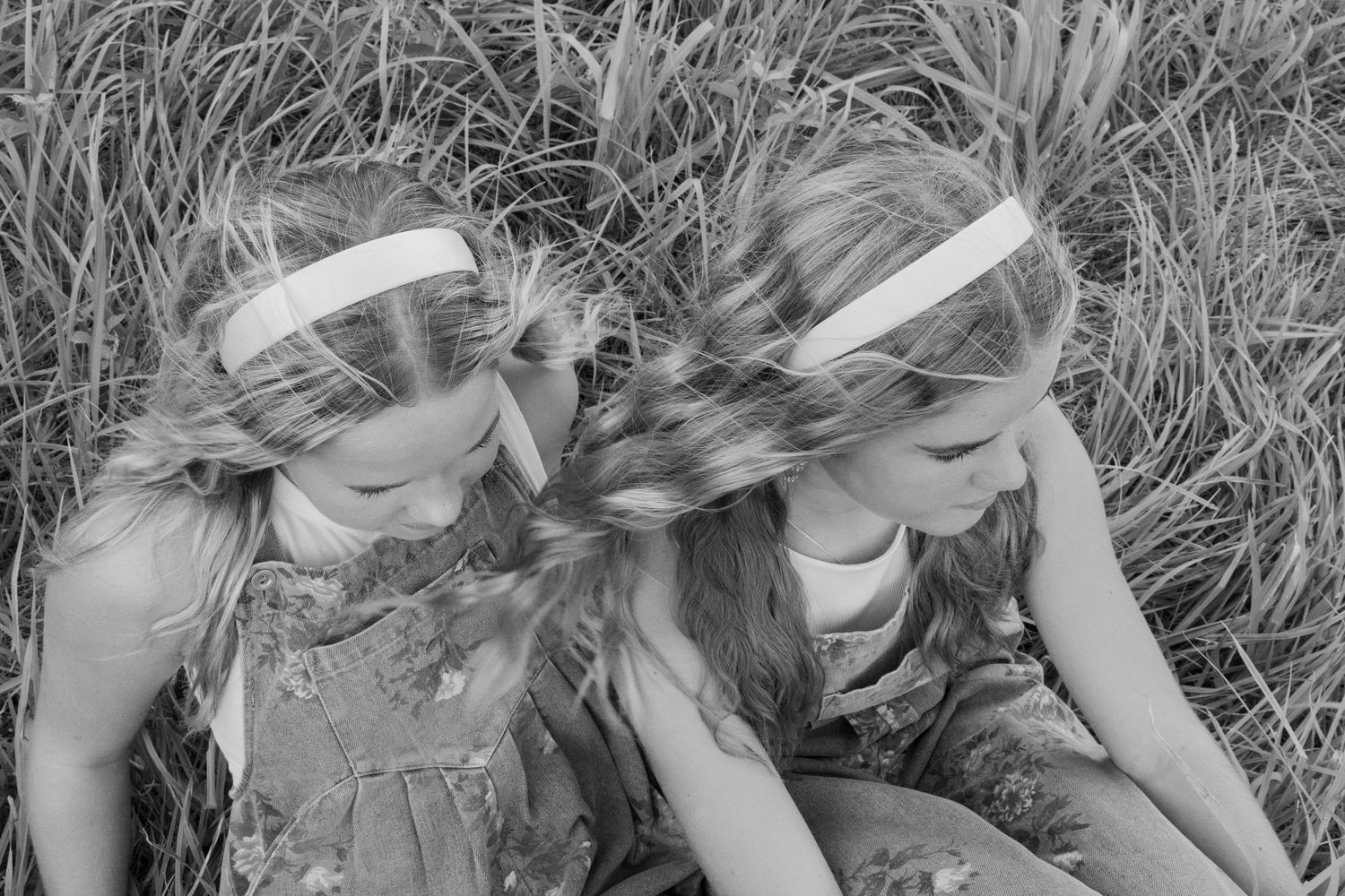 Black and white photo of two young girls with matching headbands sitting together in tall dry grass.