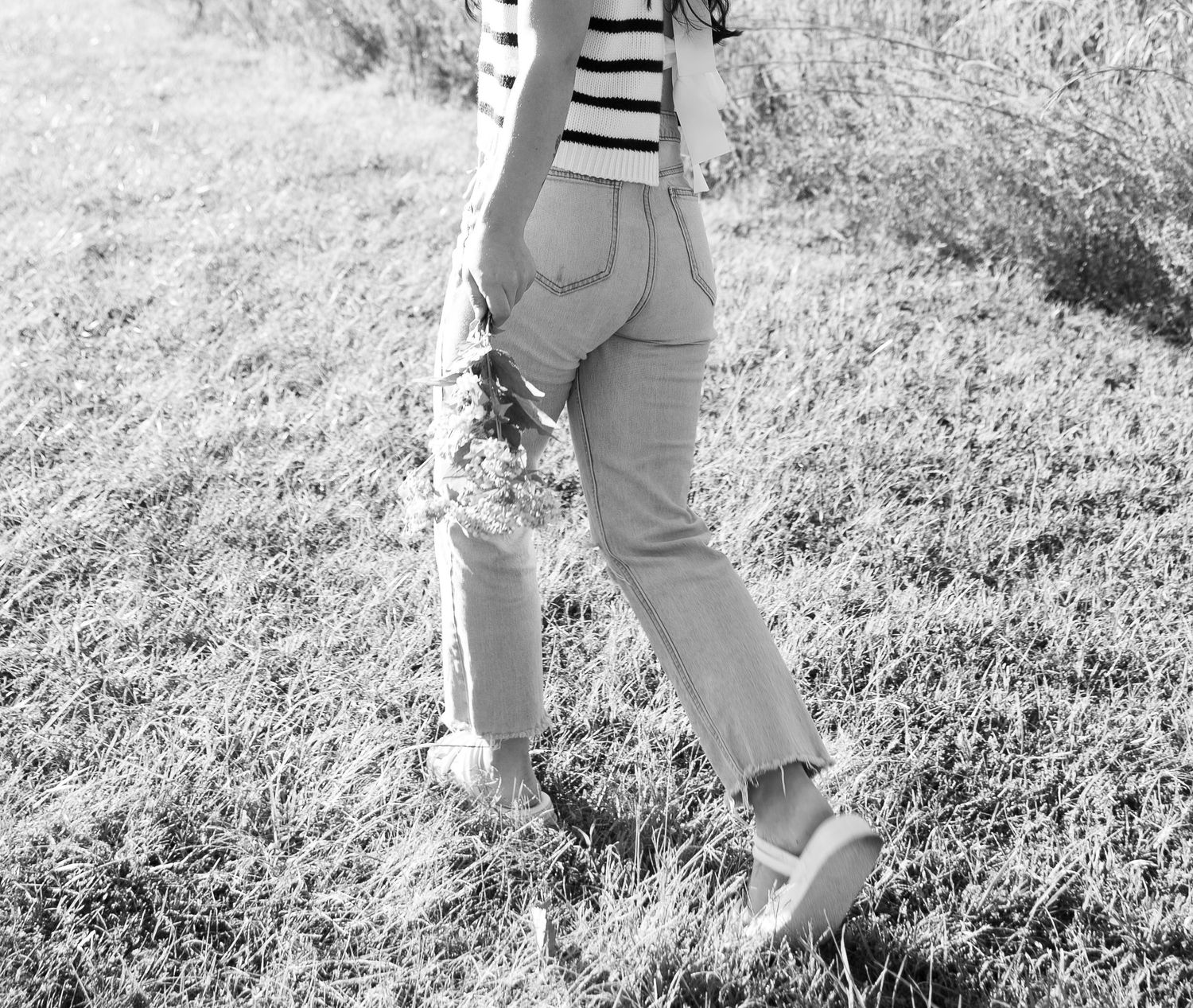 Black and white photo of a woman in striped top and jeans holding flowers while walking in a field.