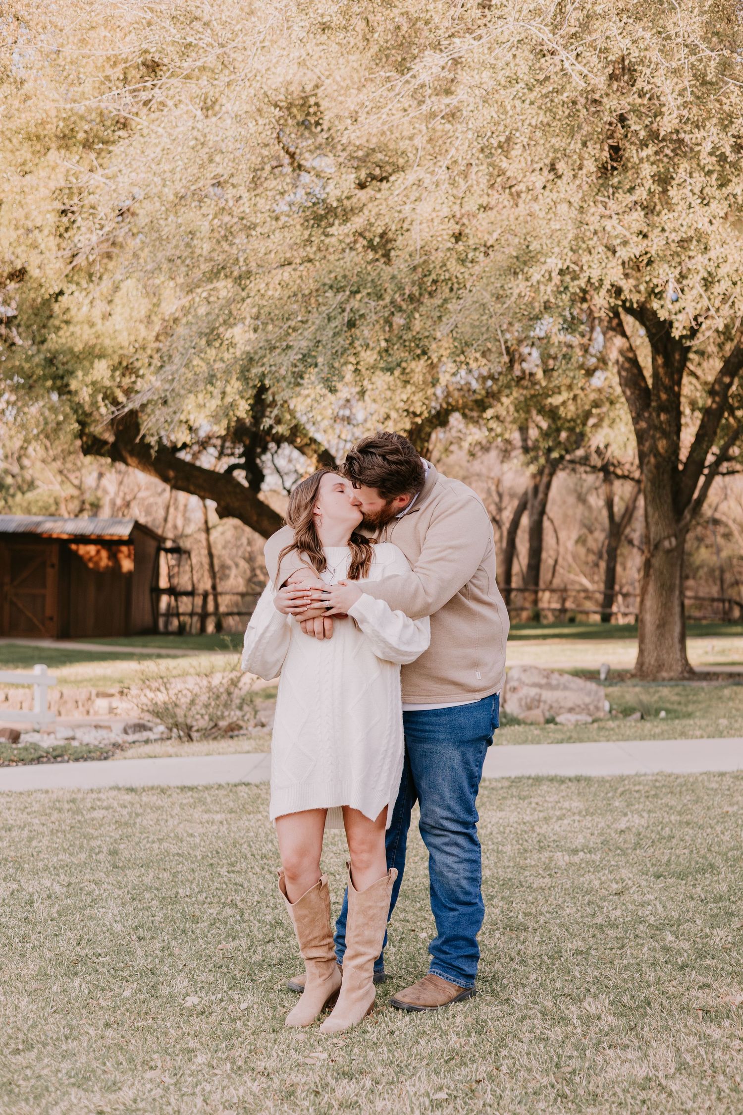 Couple embracing and kissing outdoors on green grass beneath golden autumn trees in a park setting.