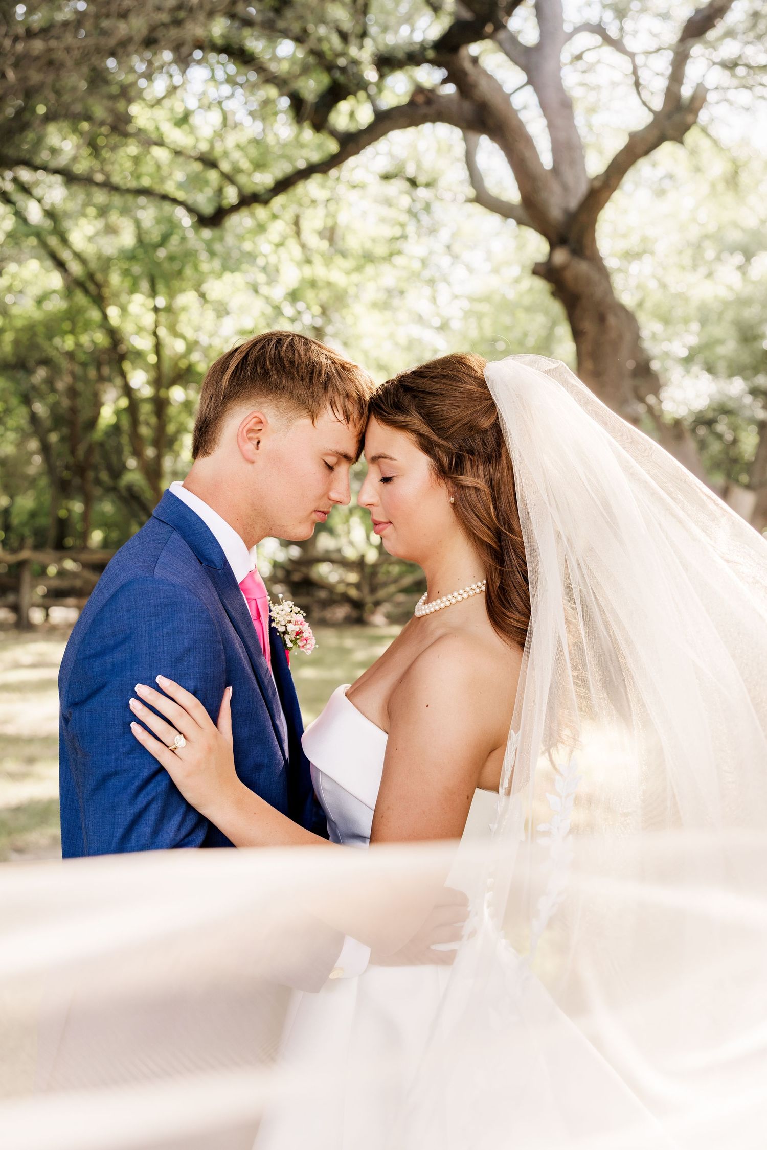 Bride and groom touching foreheads romantically outdoors under oak trees, groom in blue suit, bride in white gown with veil.