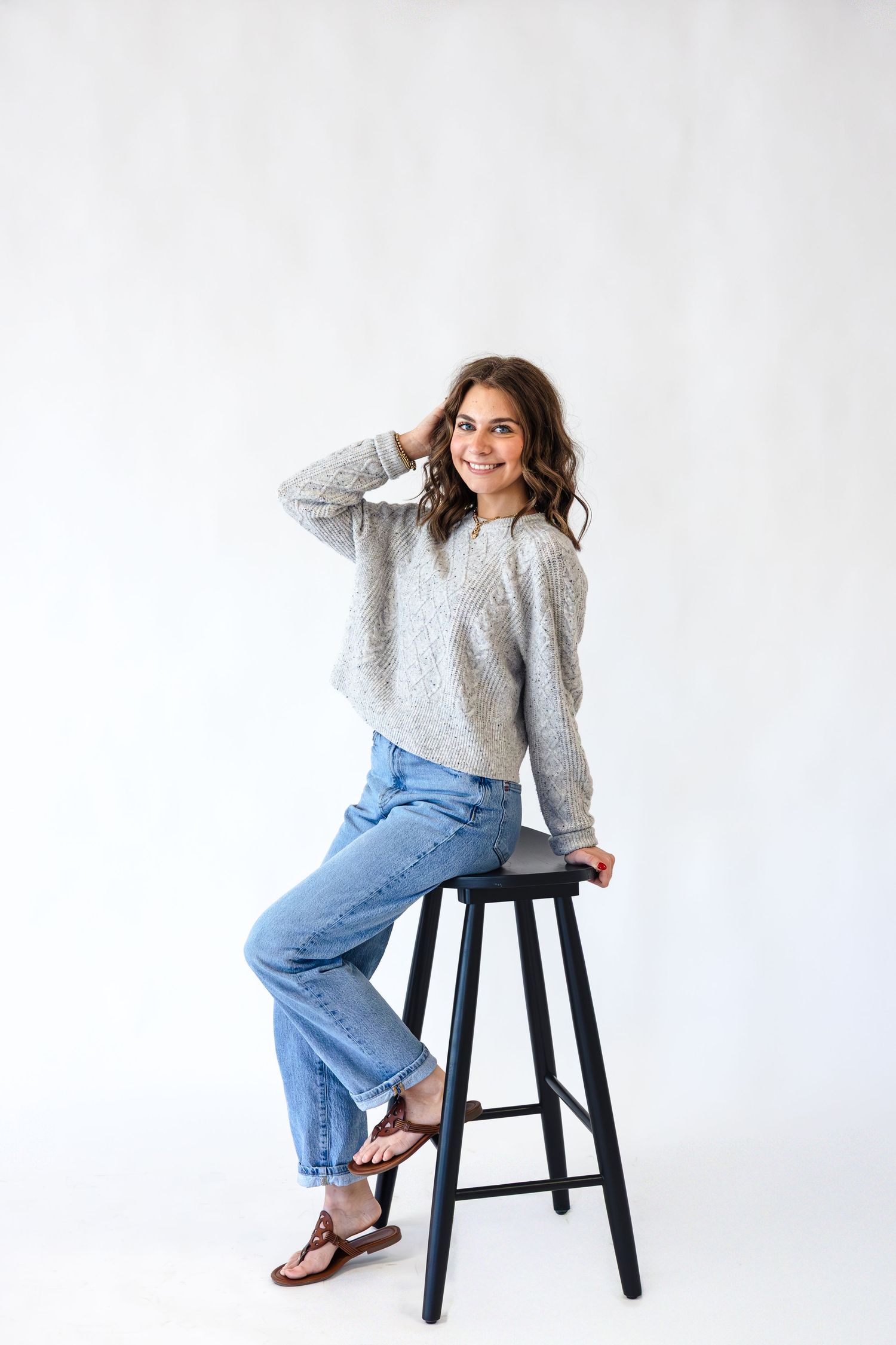 Person in a gray sweater and blue jeans poses playfully on a black stool against a white backdrop in a photo series.
