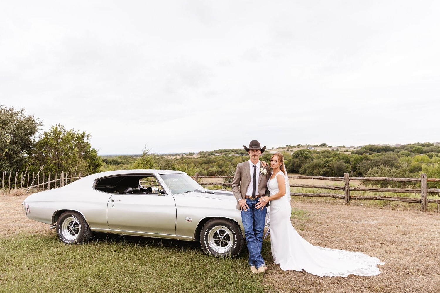 Bride and groom posing beside a classic silver muscle car on a rustic ranch with wooden fence and rolling hills.