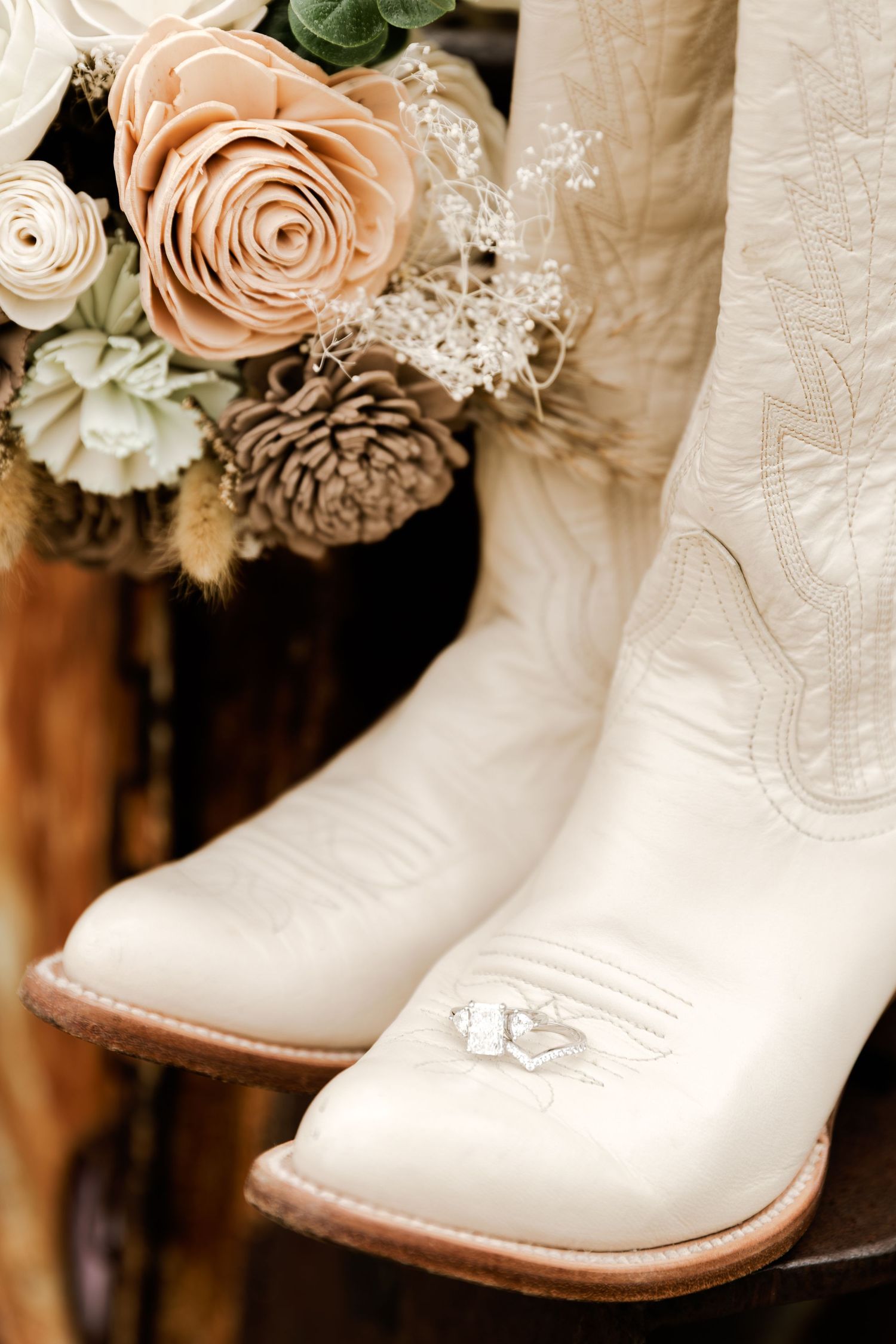 White cowboy boots with wedding rings resting beside a rustic bouquet of neutral-toned flowers and pinecones.