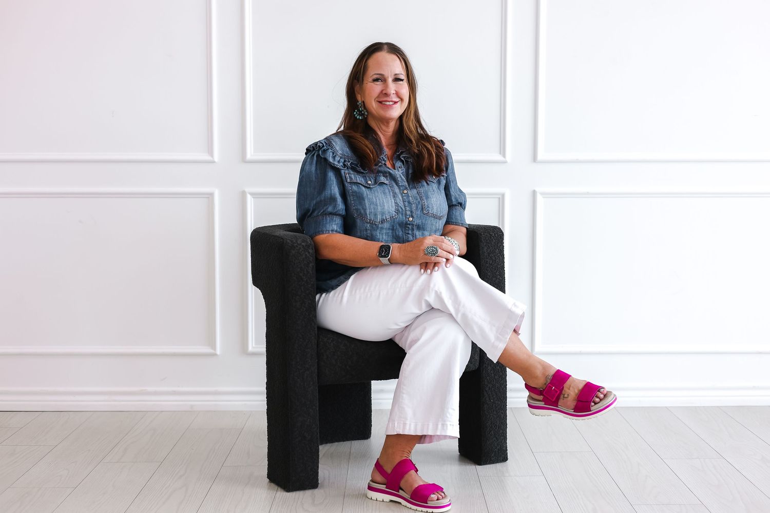 Person in denim jacket and white pants sits relaxed in black chair wearing pink sandals.