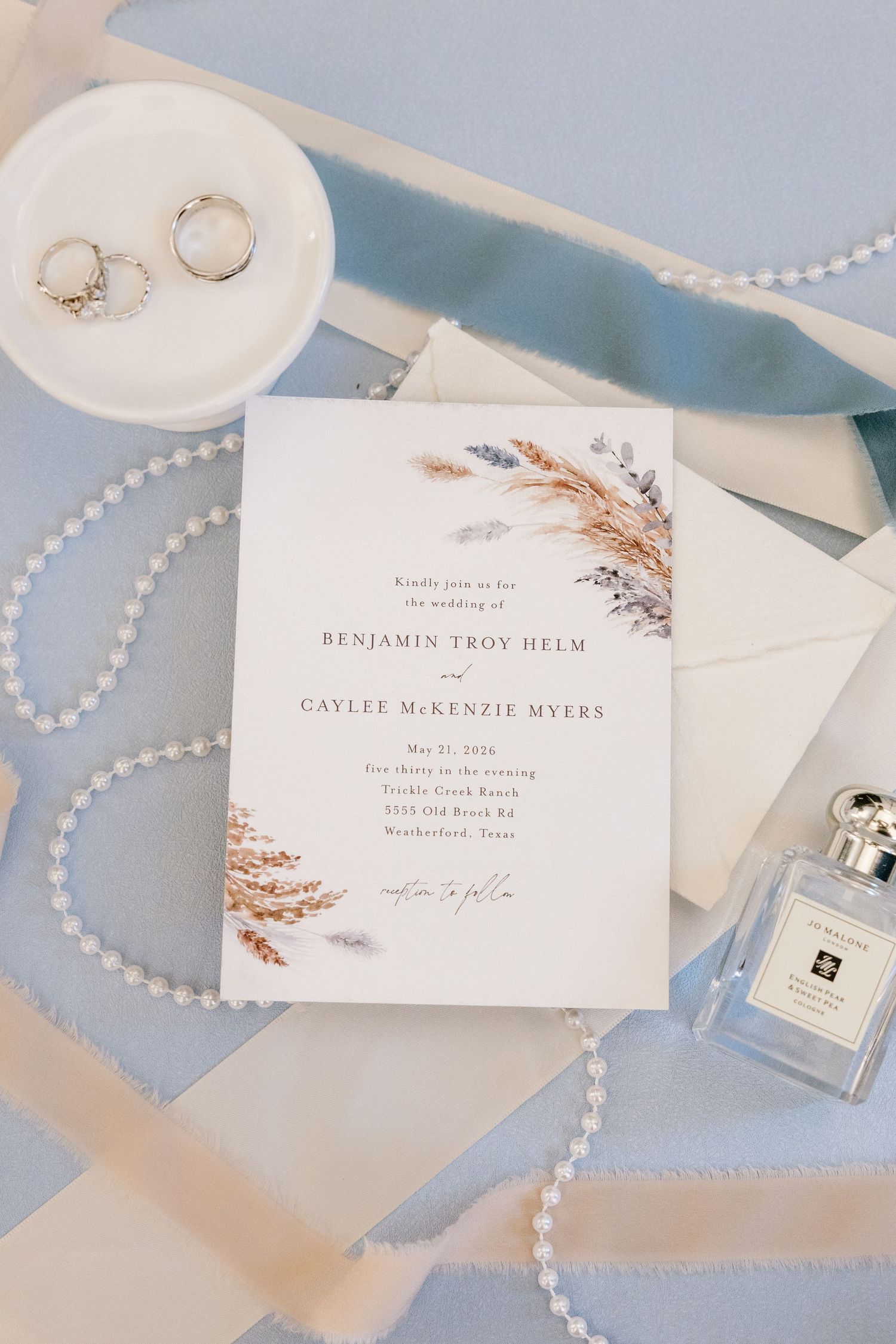 Elegant wedding invitation for Benjamin Troy Helm and Caylee McKenzie Myers with pampas grass, pearl necklace, rings, and perfume on blue.