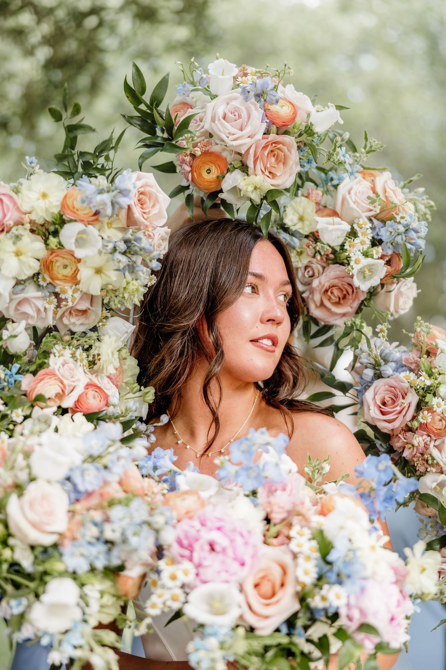 Young woman surrounded by lush pastel floral arrangements featuring roses, ranunculus, and blue blooms in an elegant outdoor setting.