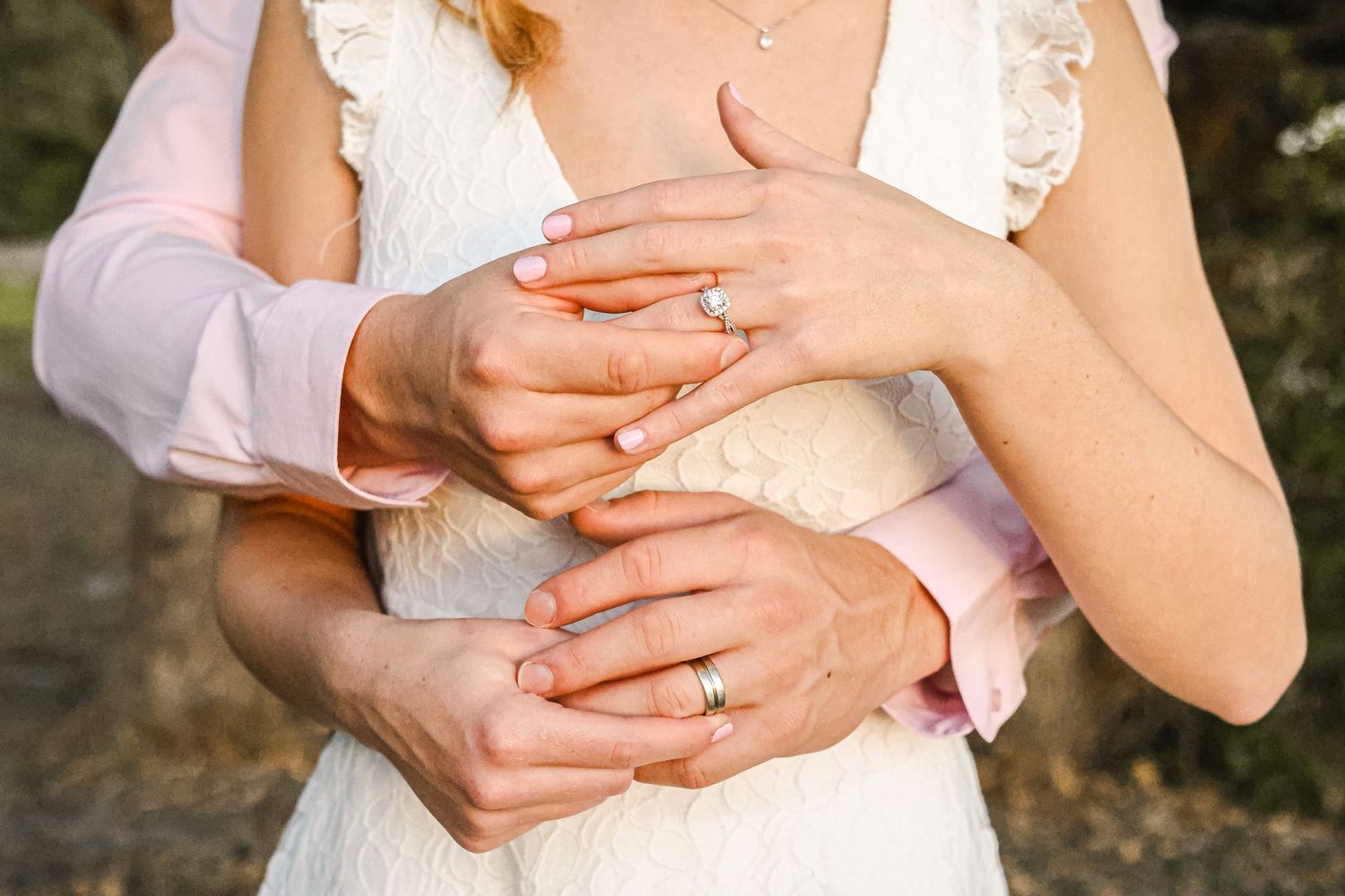Close up shot of hands holding each other with wedding rings against a white dress in an outdoor setting.