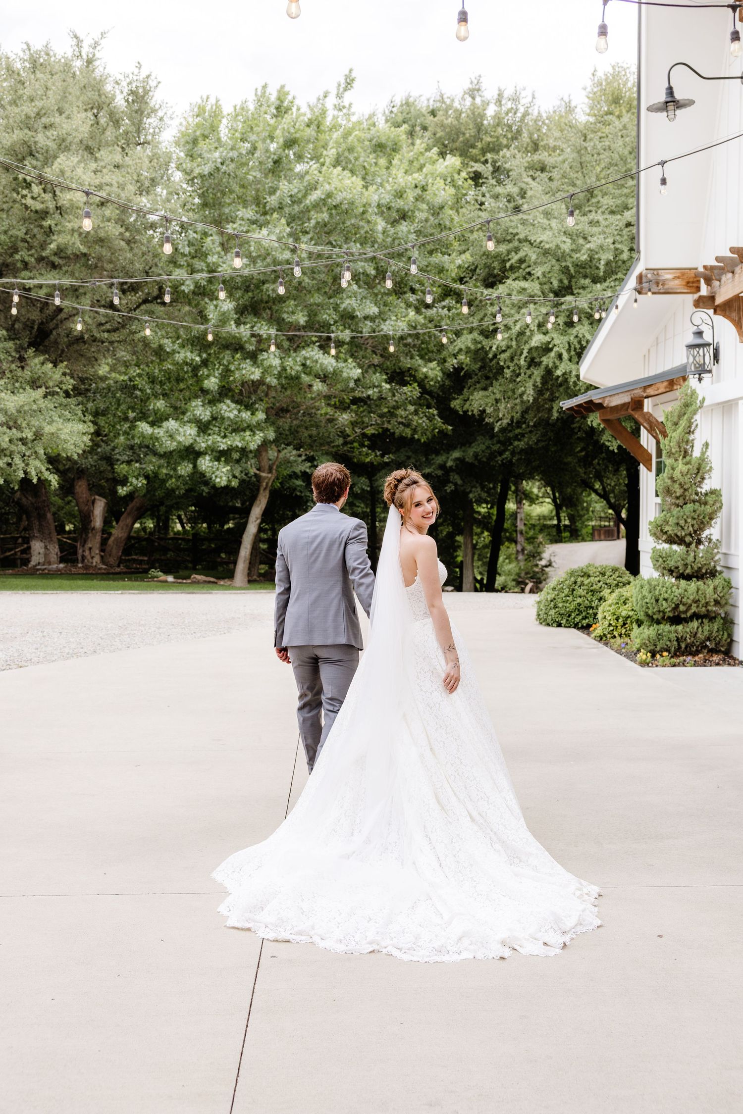 Bride in elegant lace gown with train looks back while groom in gray suit walks ahead at outdoor wedding venue.
