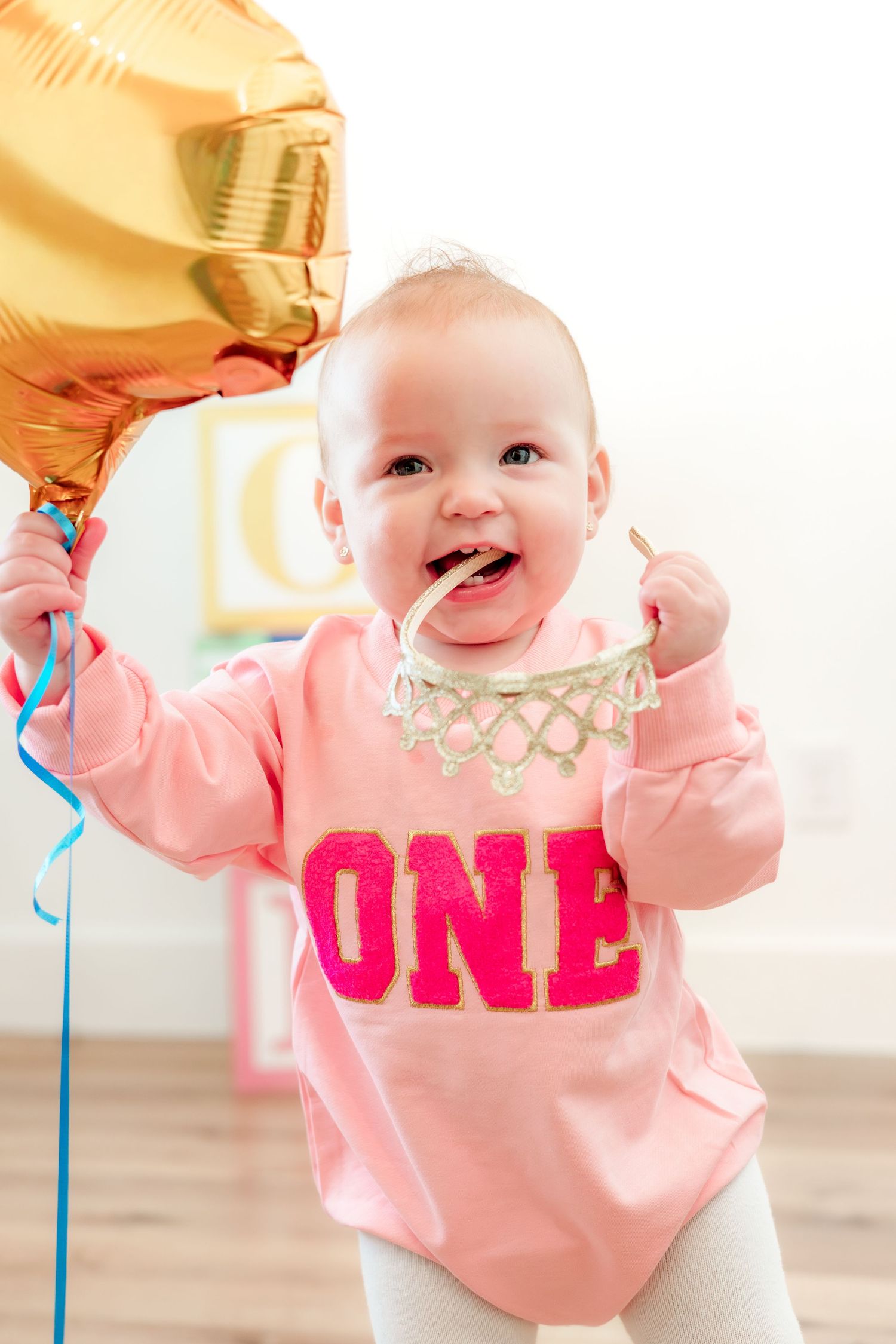 Baby girl in pink ONE sweatshirt holds gold balloon celebrating her first birthday party.