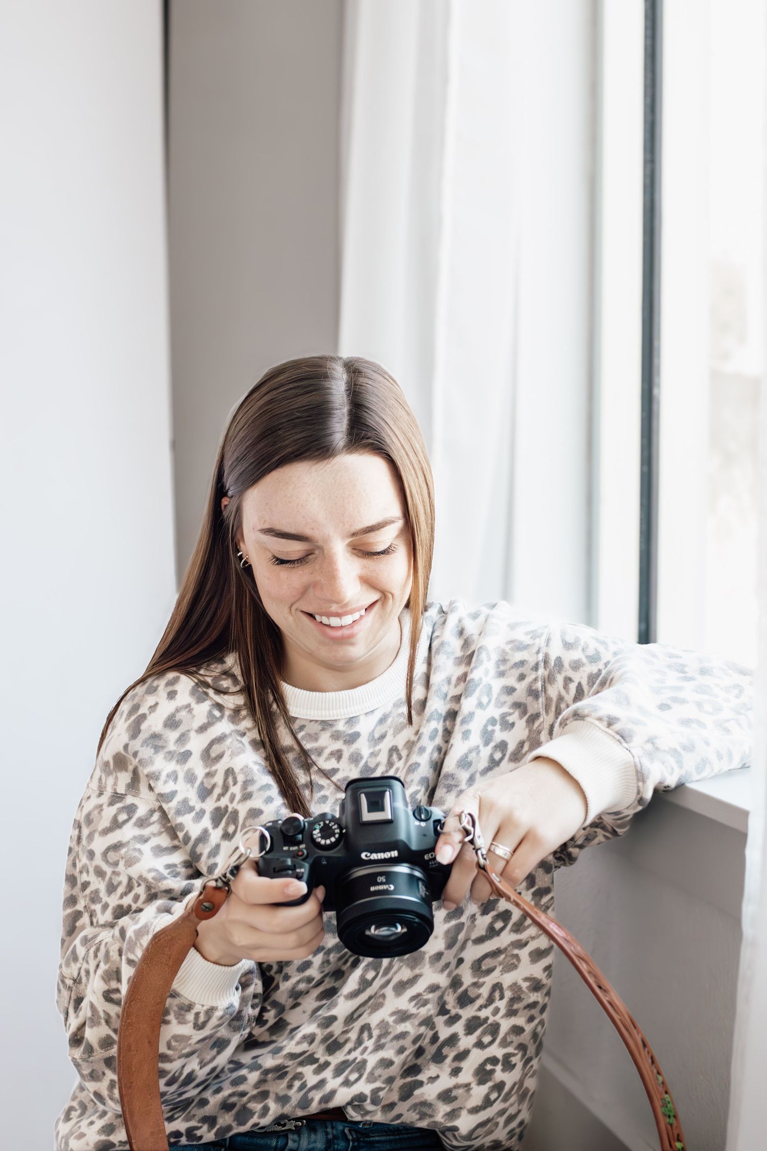 Smiling woman in leopard print sweater reviewing photos on Canon DSLR camera near bright window.
