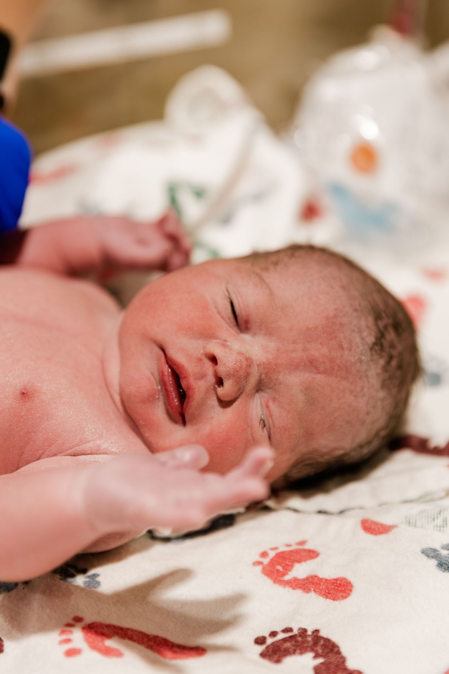 Newborn baby lying on a colorful footprint blanket in a hospital nursery, eyes closed, shortly after birth.