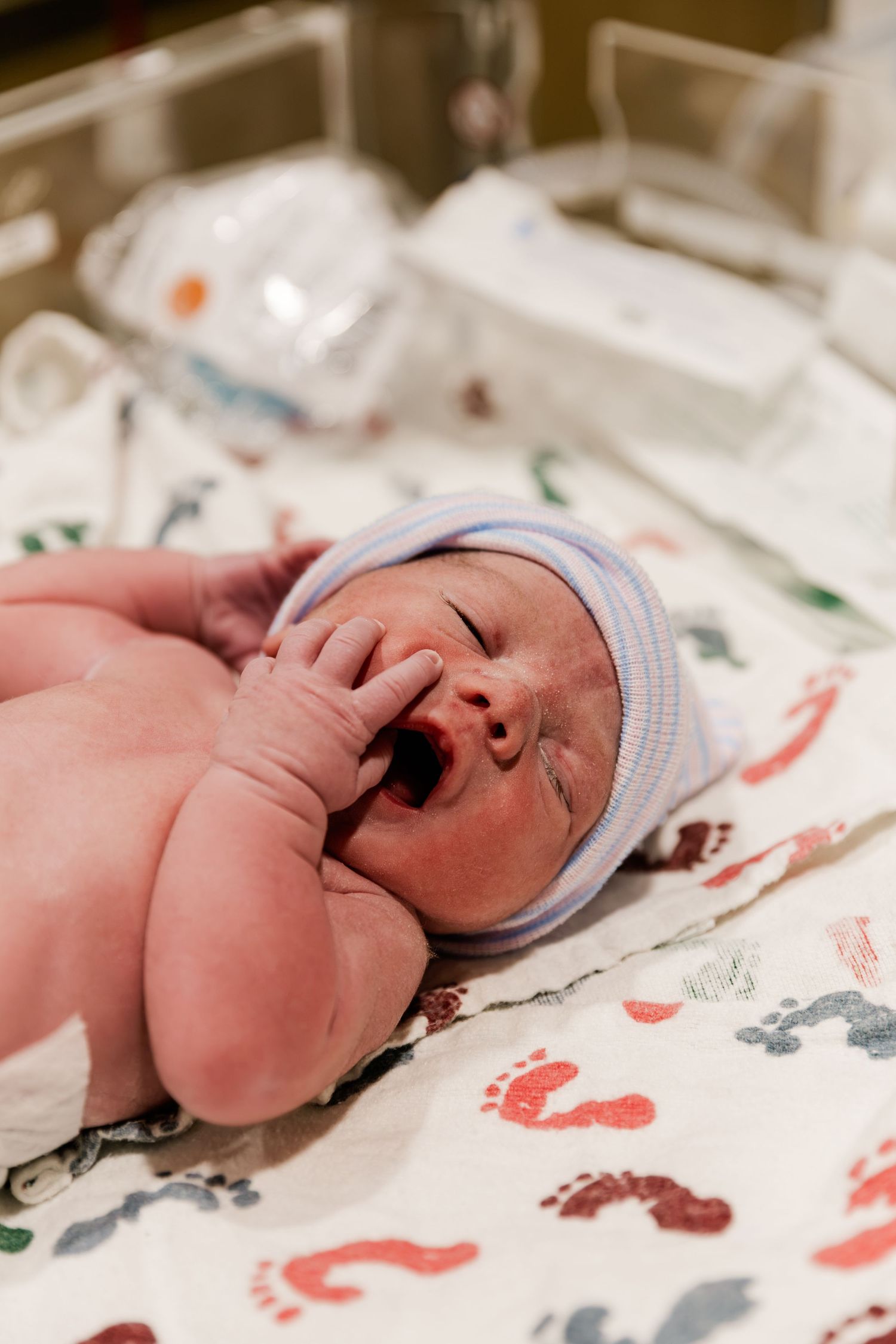 Newborn baby yawning in hospital nursery, wearing striped hat, lying on colorful footprint blanket.