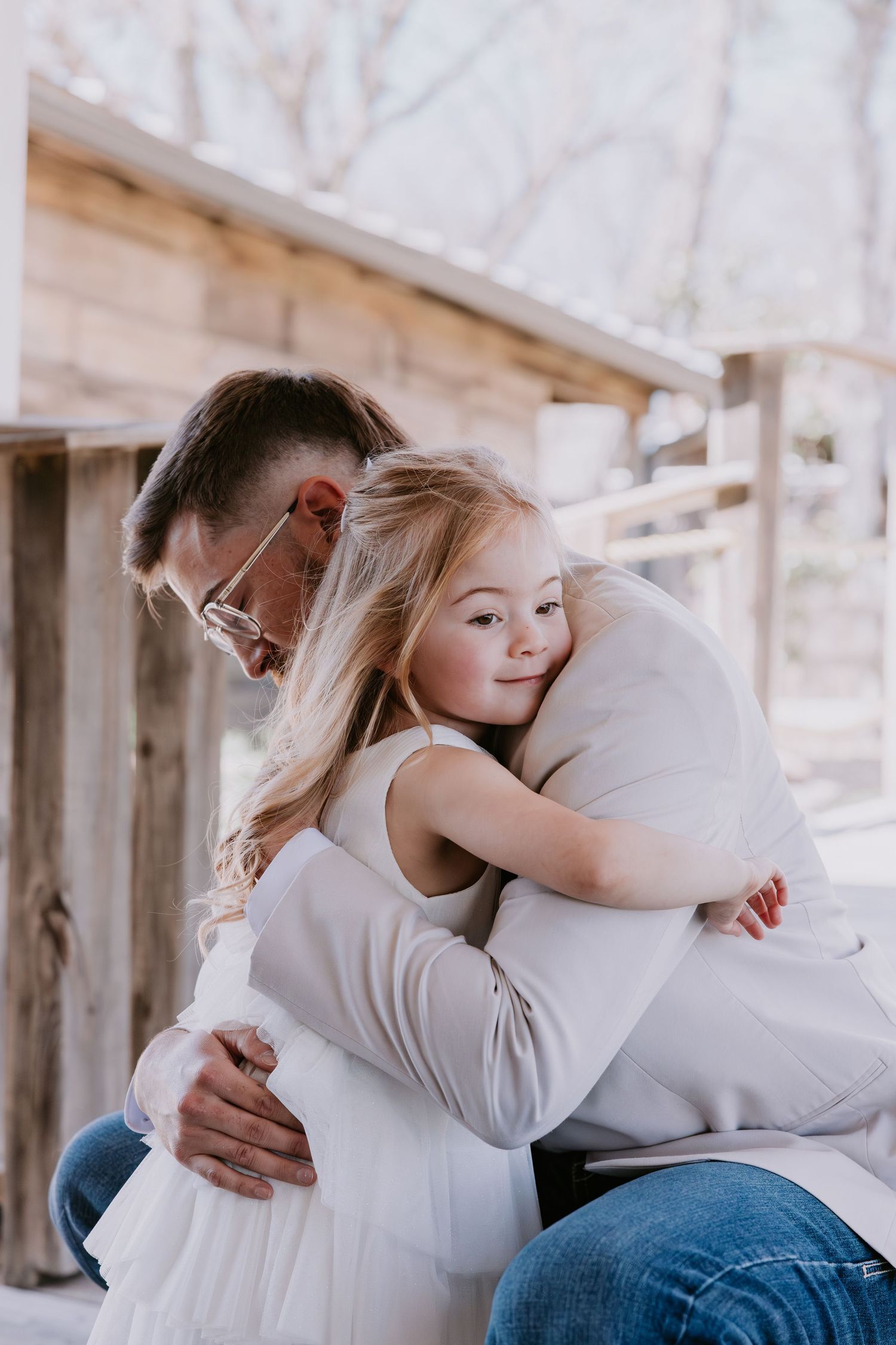 A young girl with blonde hair hugs a man wearing glasses outdoors near a rustic wooden structure.
