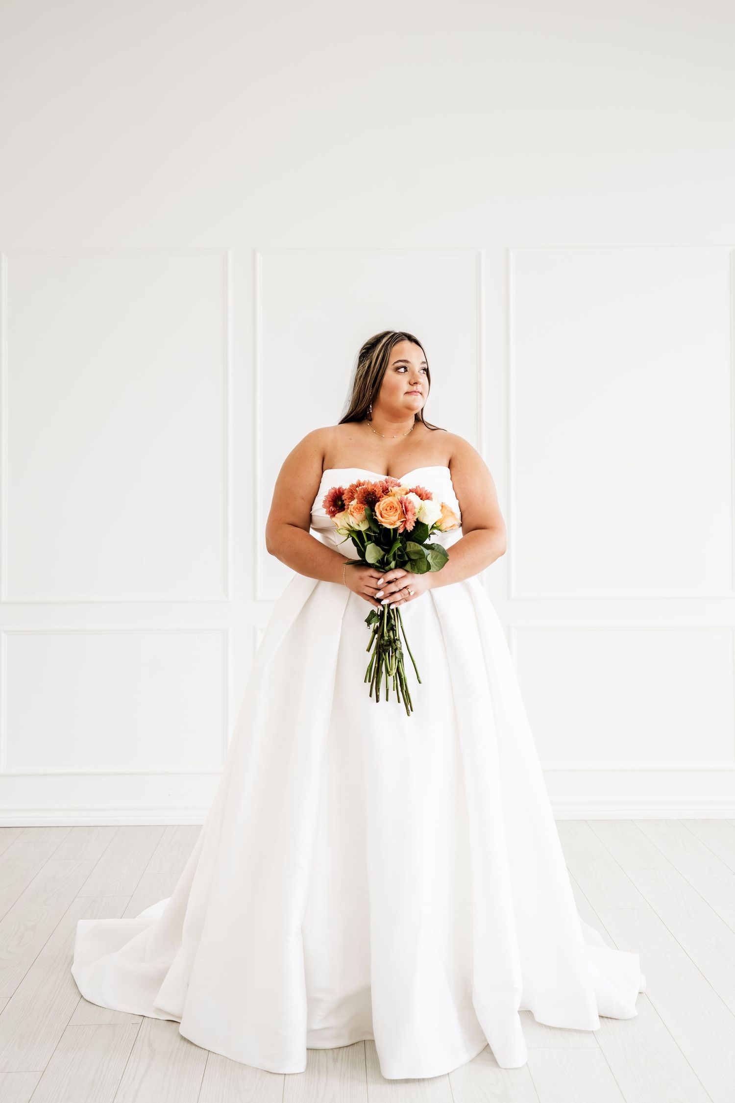 Bride in strapless white ballgown holding orange and pink rose bouquet against white wall.