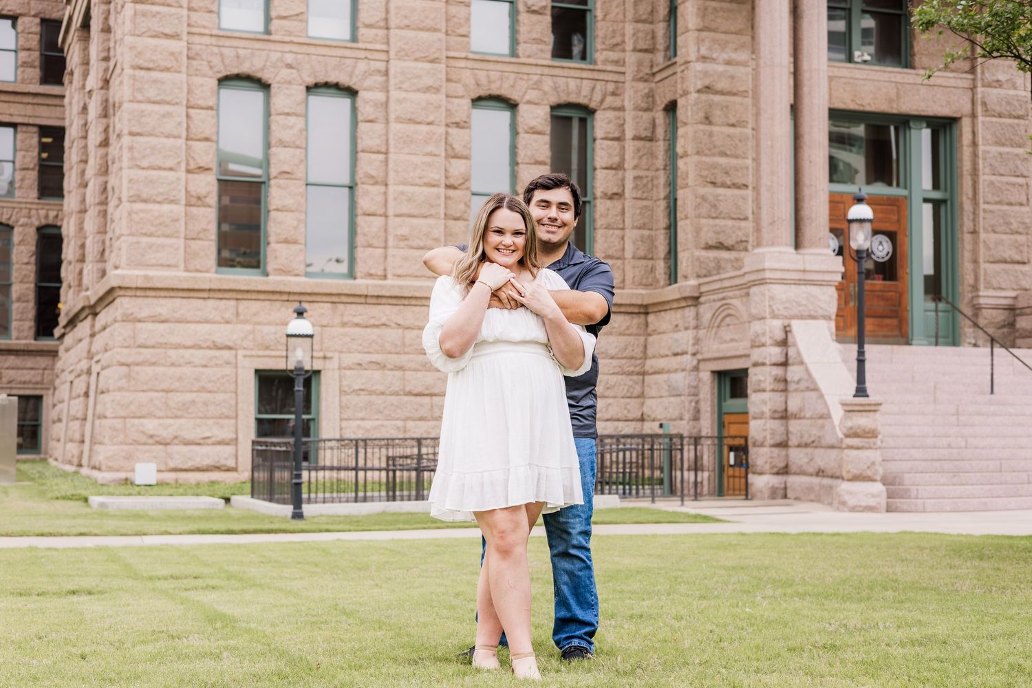 Couple embracing and smiling in front of a historic brick building on a green lawn.