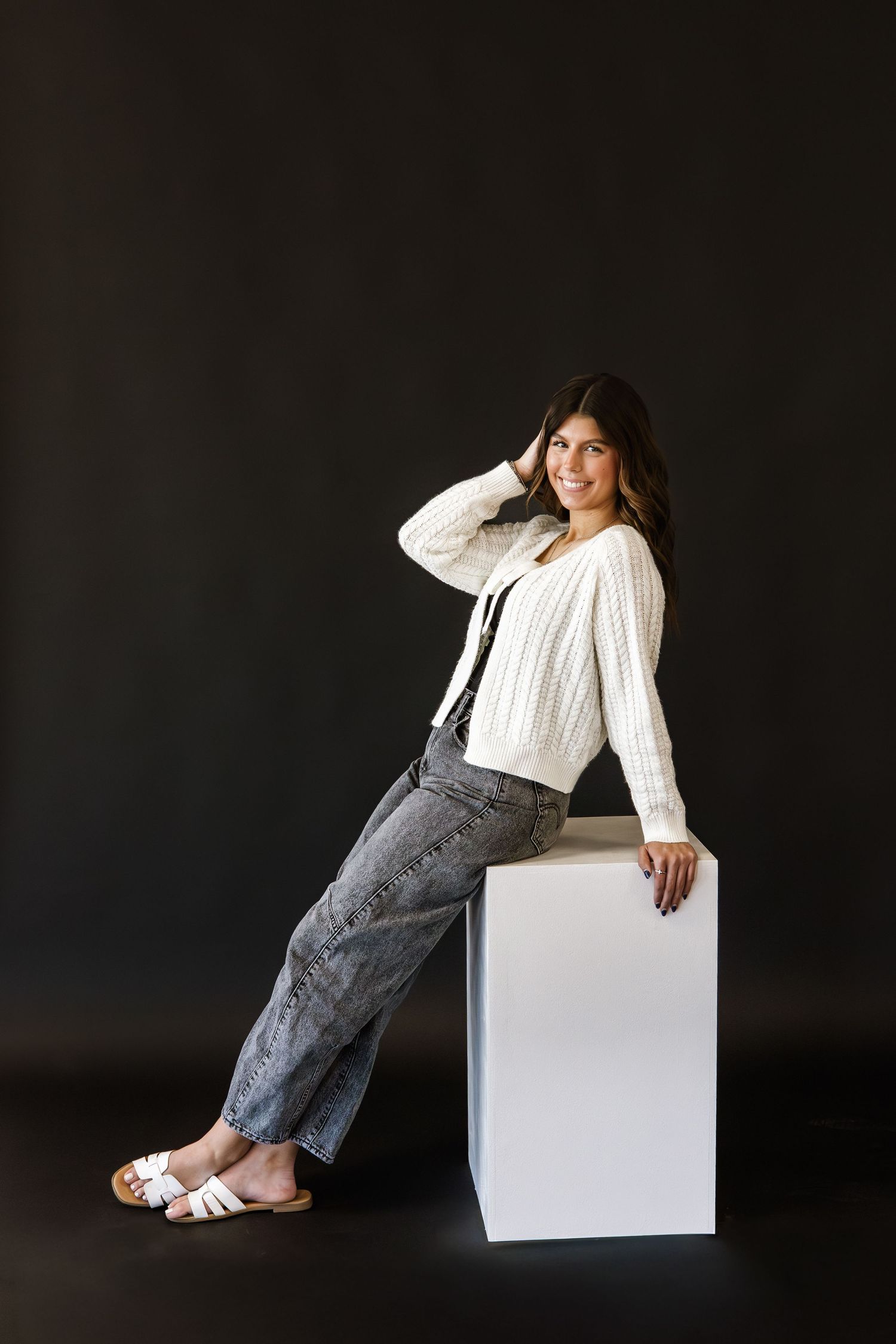 Woman in white cable-knit sweater and gray jeans leaning on white cube, smiling against dark background.