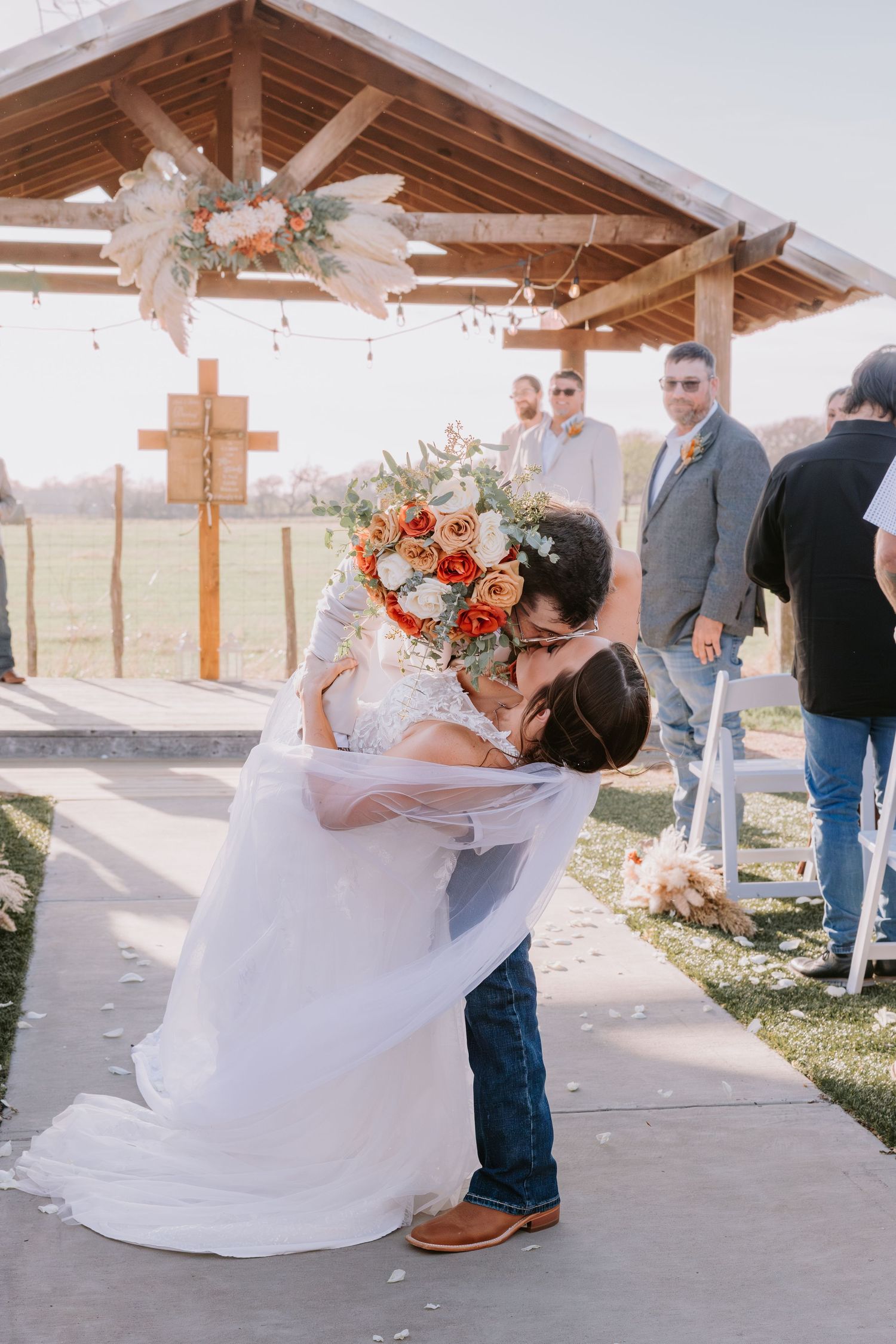 Bride and groom share a romantic dip kiss at outdoor rustic wedding ceremony with floral arch and guests watching.