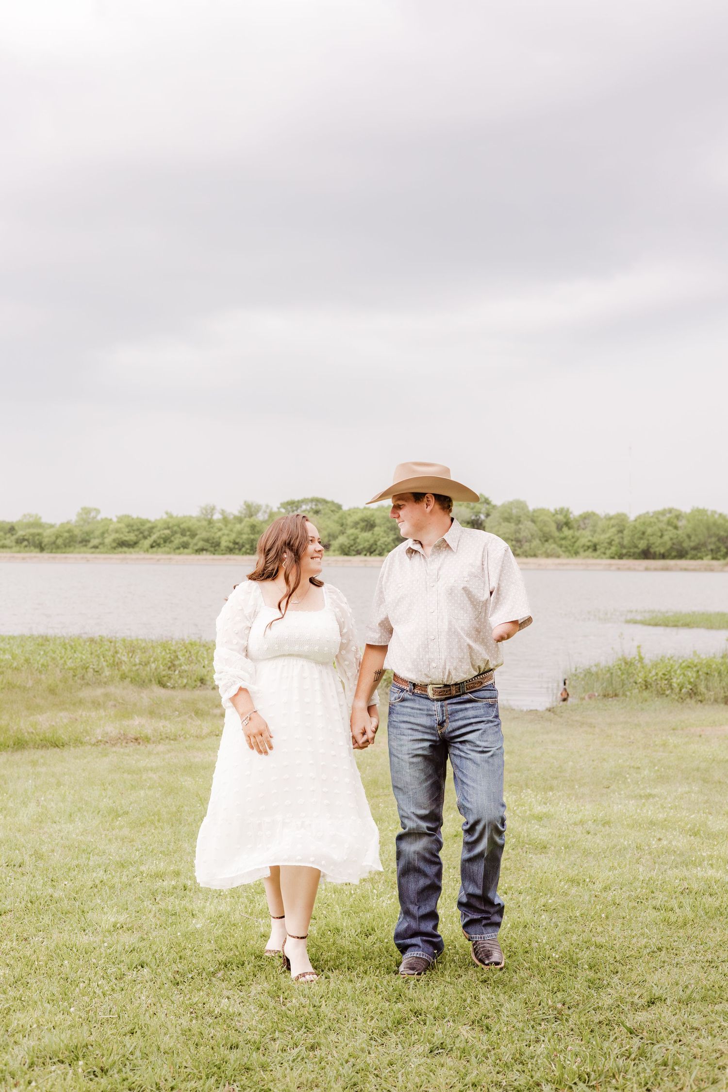 Couple holding hands walking by a lake, woman in white dress, man in cowboy hat and jeans, outdoor portrait.