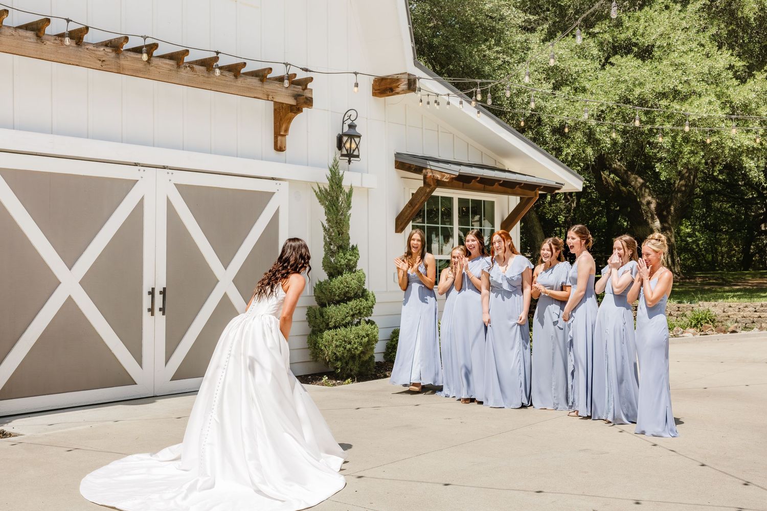 Bride in white gown reveals look to bridesmaids in lavender dresses outside a white barn wedding venue.