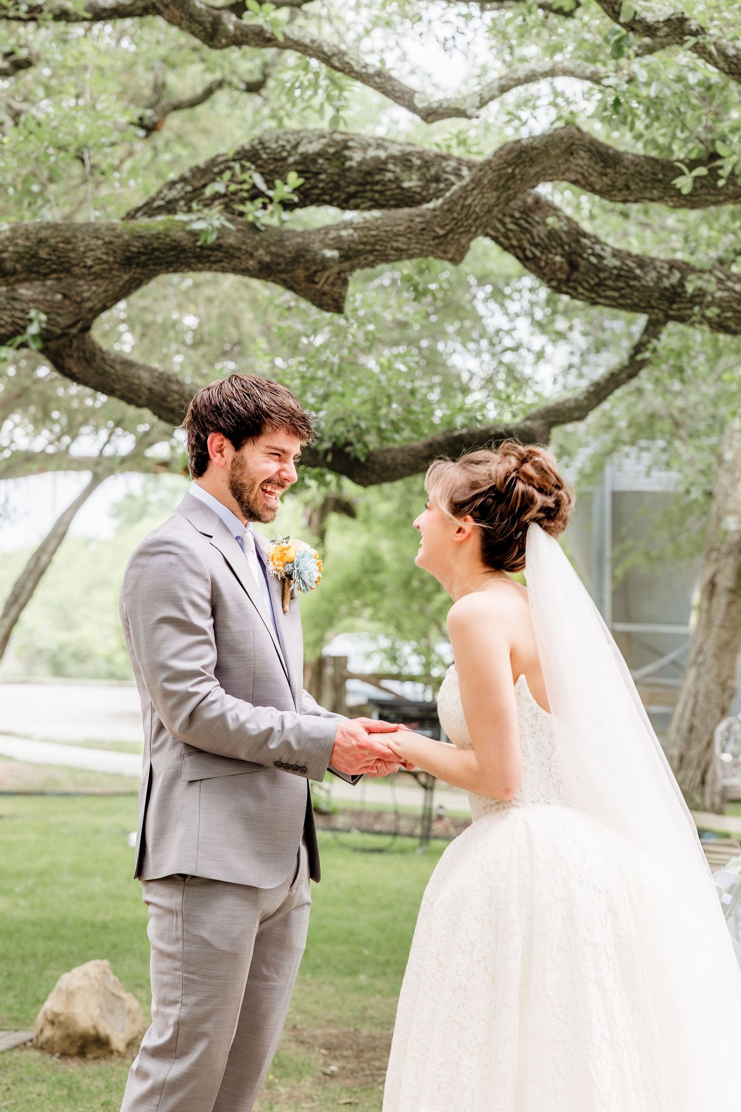Smiling groom in gray suit holds bride's hands outdoors under large oak tree during romantic wedding first look.