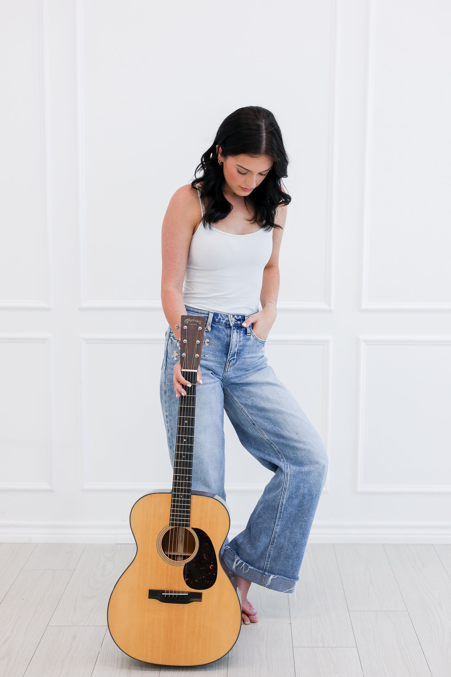 Person in white tank top and jeans poses with acoustic guitar against white wall.