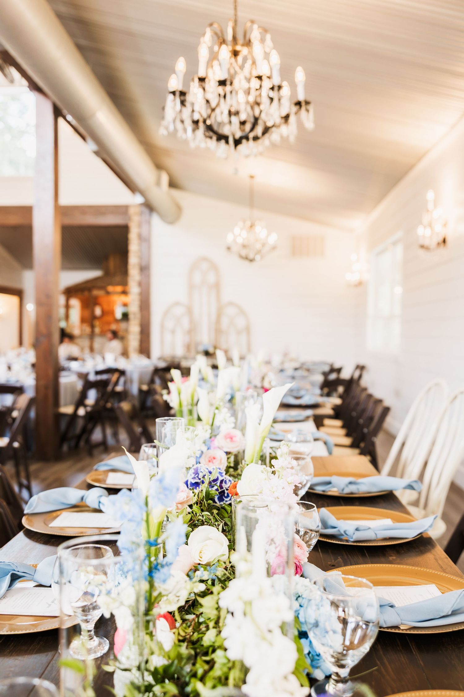 Elegant wedding reception table with floral centerpieces, blue napkins, gold chargers, and crystal chandeliers overhead.