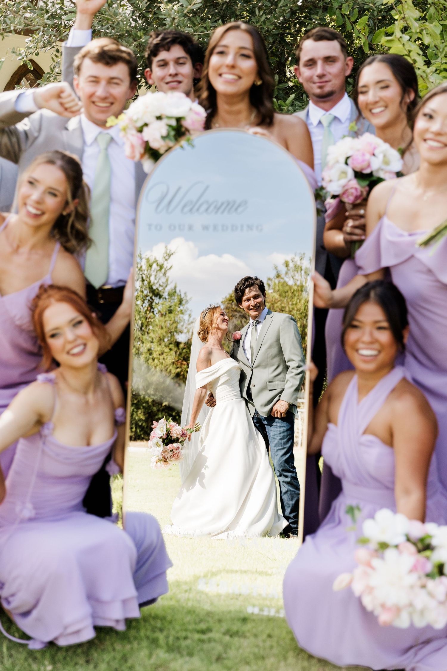 Wedding party in lavender dresses poses around a decorative mirror reflecting the bride and groom at an outdoor ceremony.