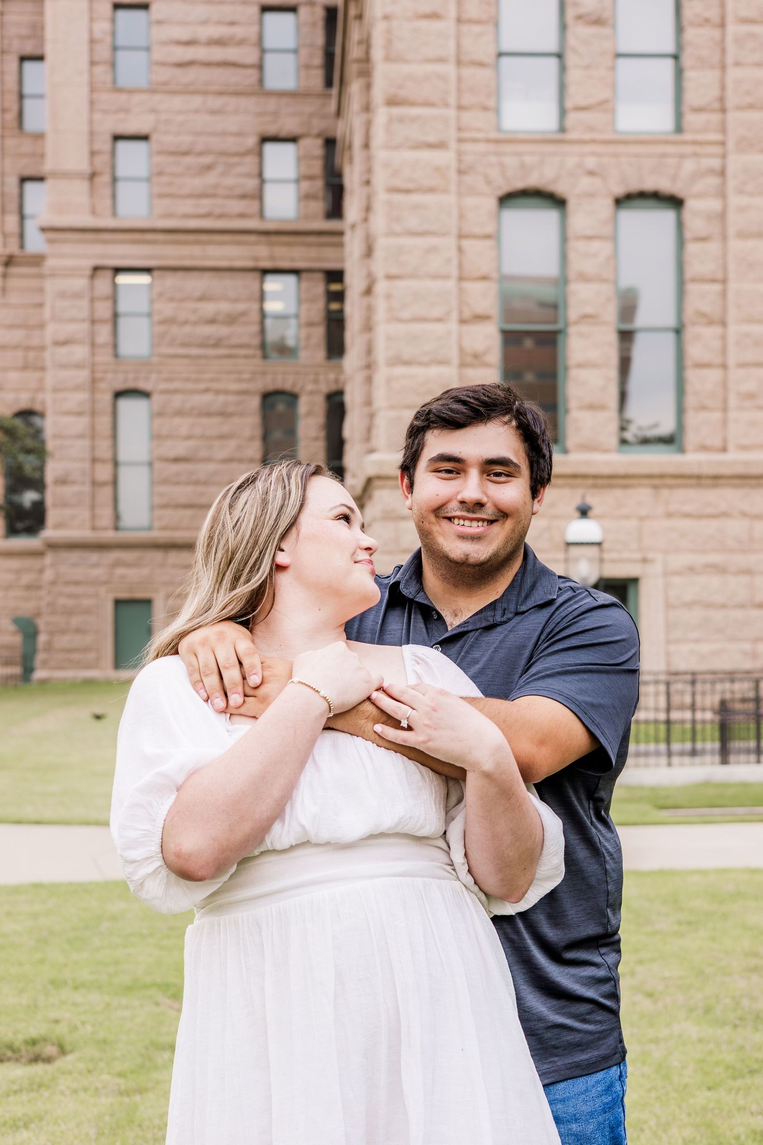 A couple embraces and smiles in front of a brick building, woman in white dress, man in navy shirt.