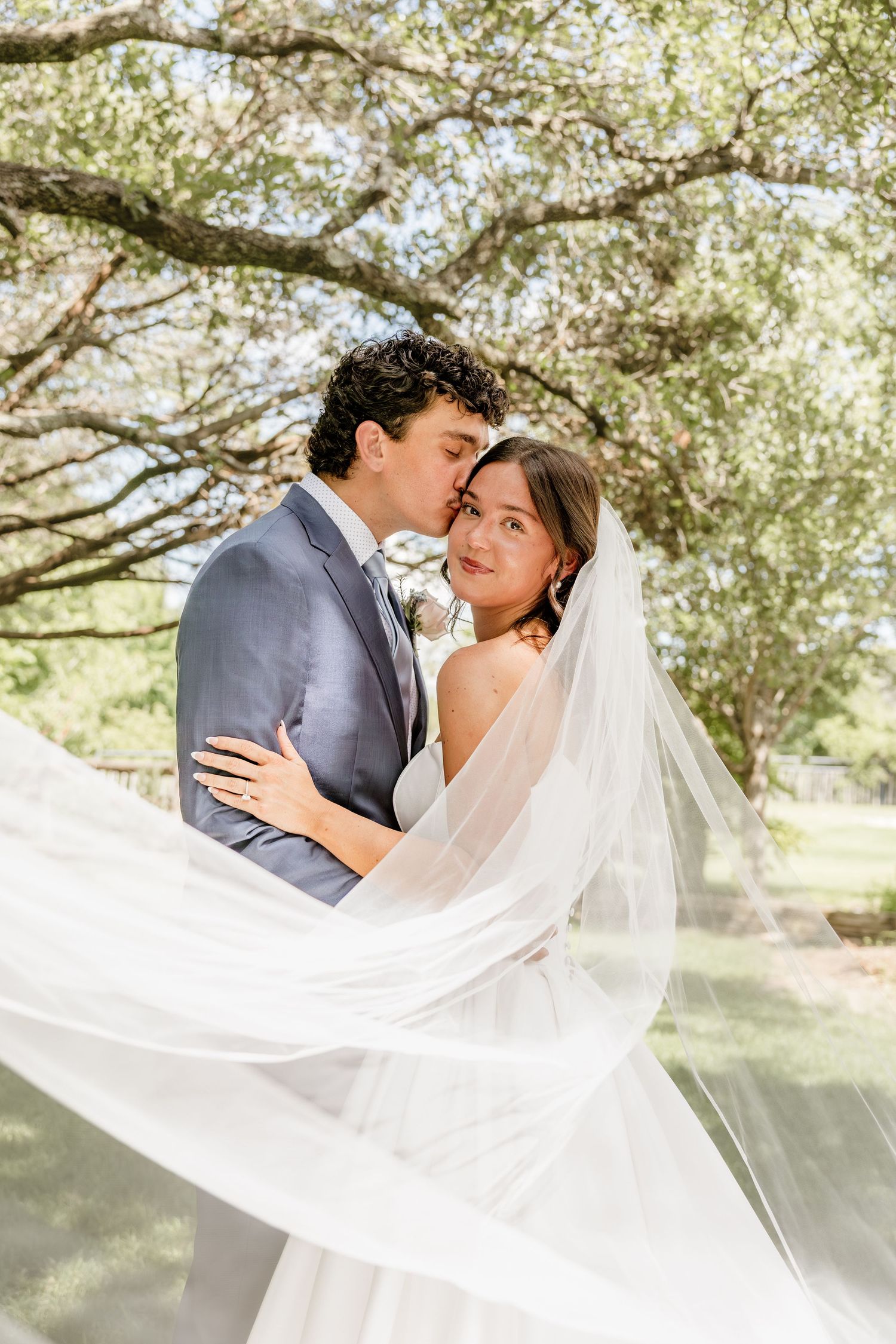 Bride and groom share a romantic kiss under oak trees, with flowing veil billowing in the breeze on their wedding day.