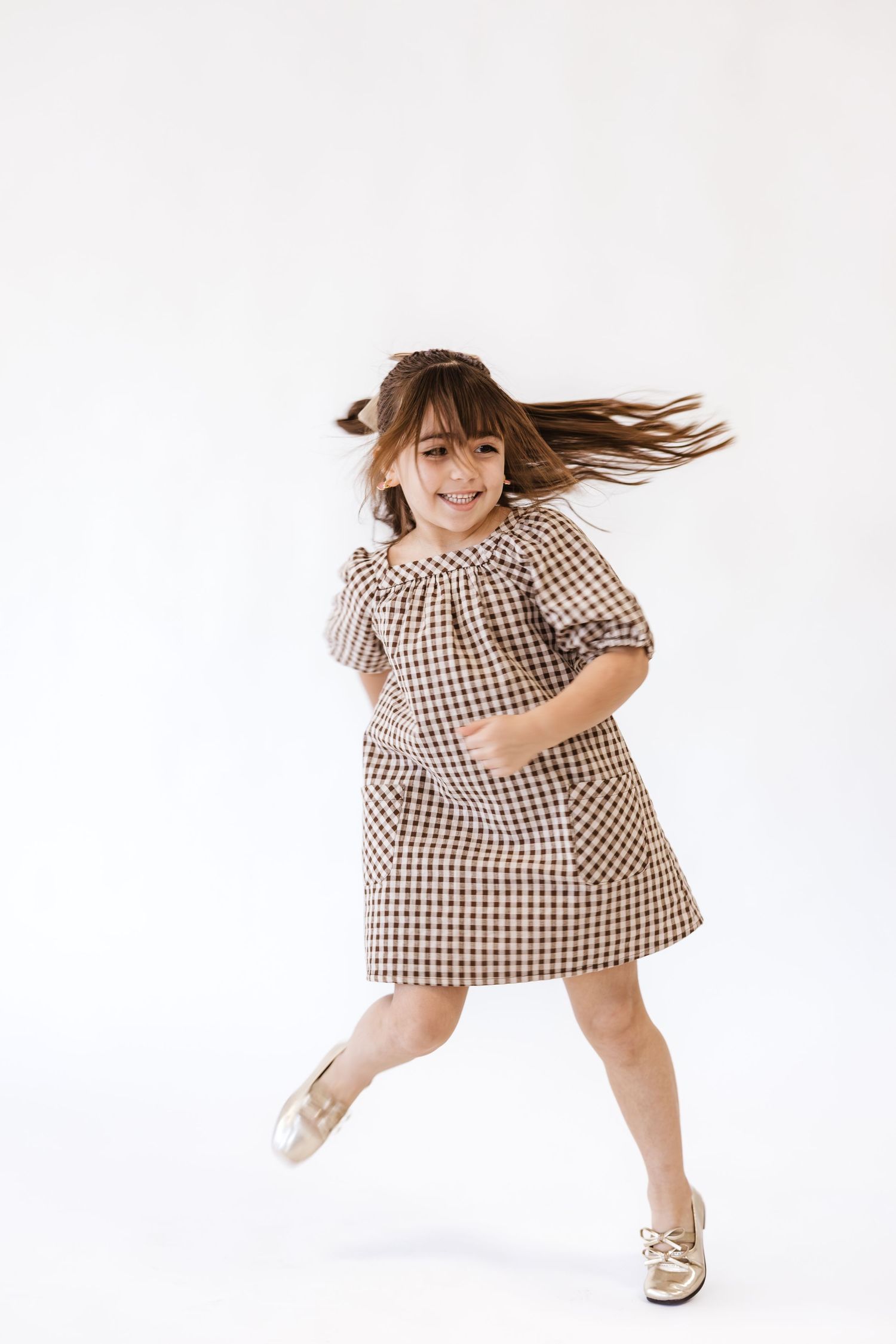 Young girl in checkered dress dancing joyfully with hair flowing, wearing gold shoes on white background.