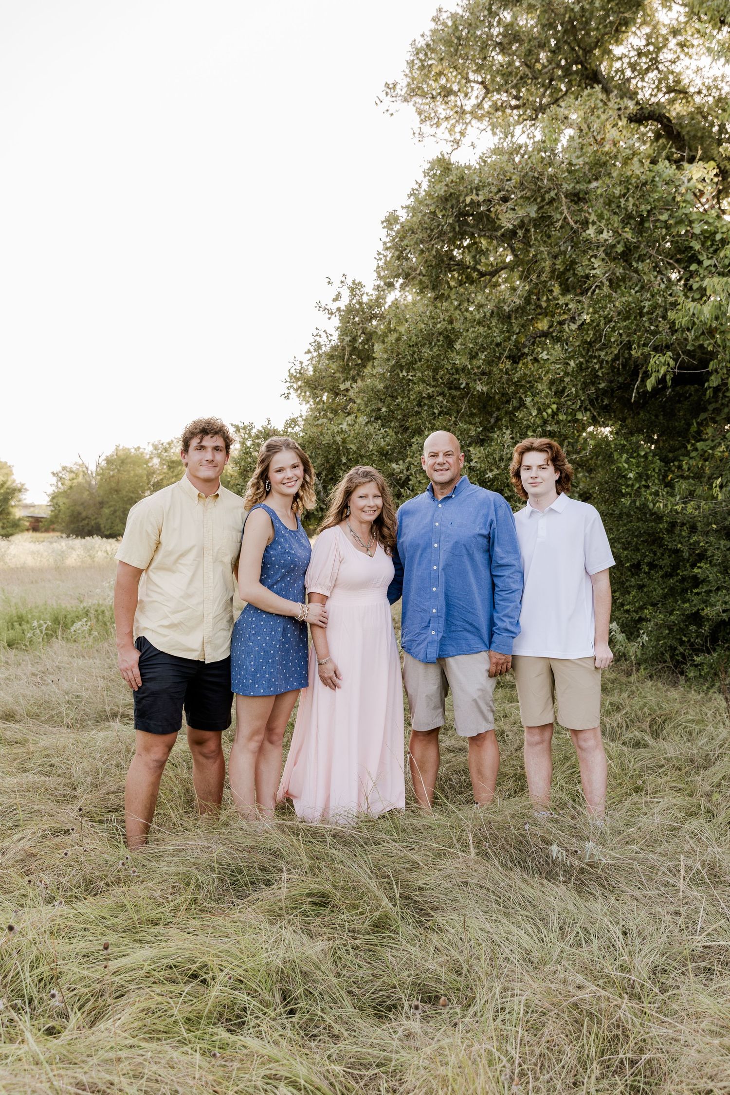 Family of five posing outdoors in a grassy field with trees, wearing casual summer attire for a portrait.