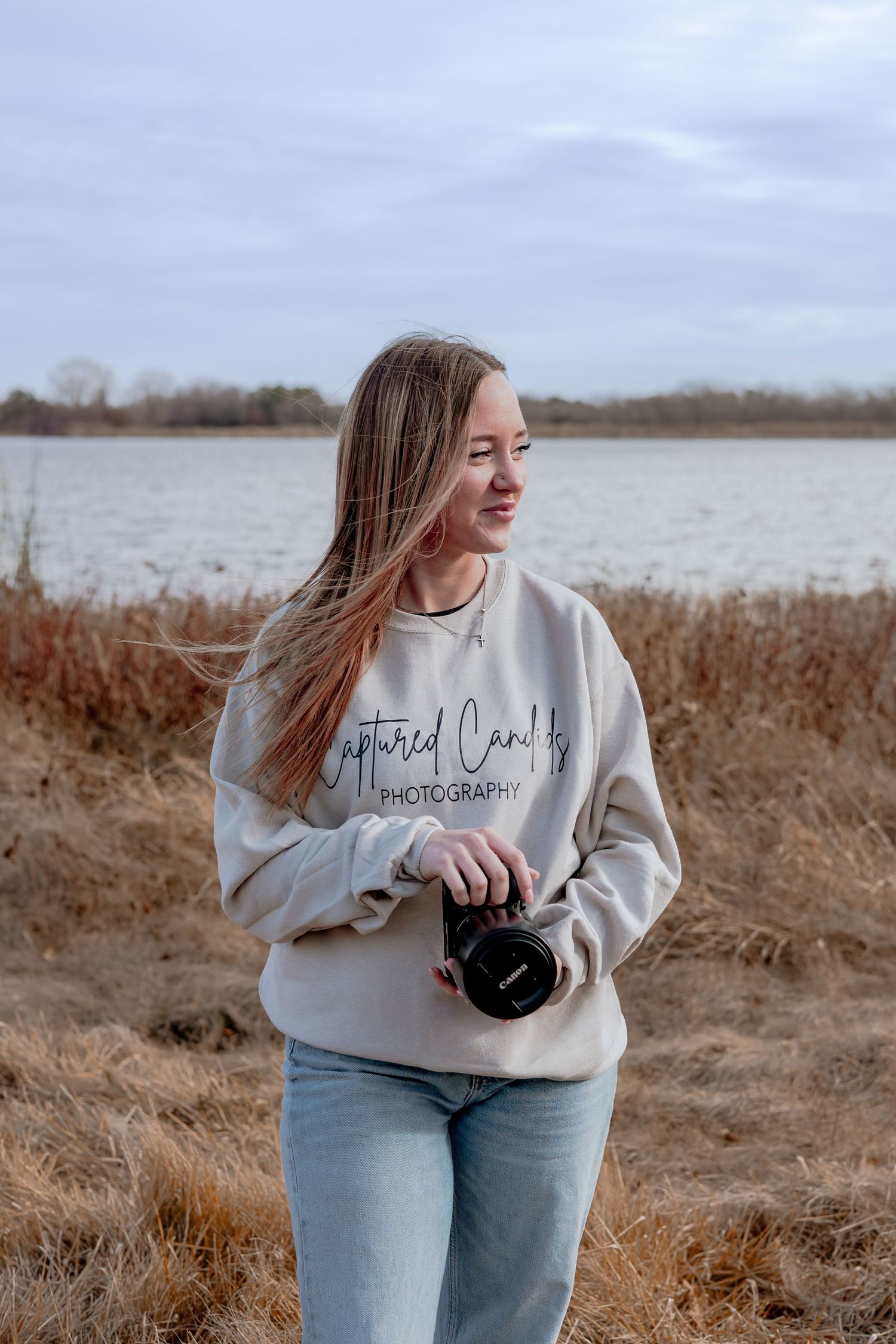 Young female photographer holding a camera outdoors near a lake, wearing a beige photography sweatshirt and jeans.