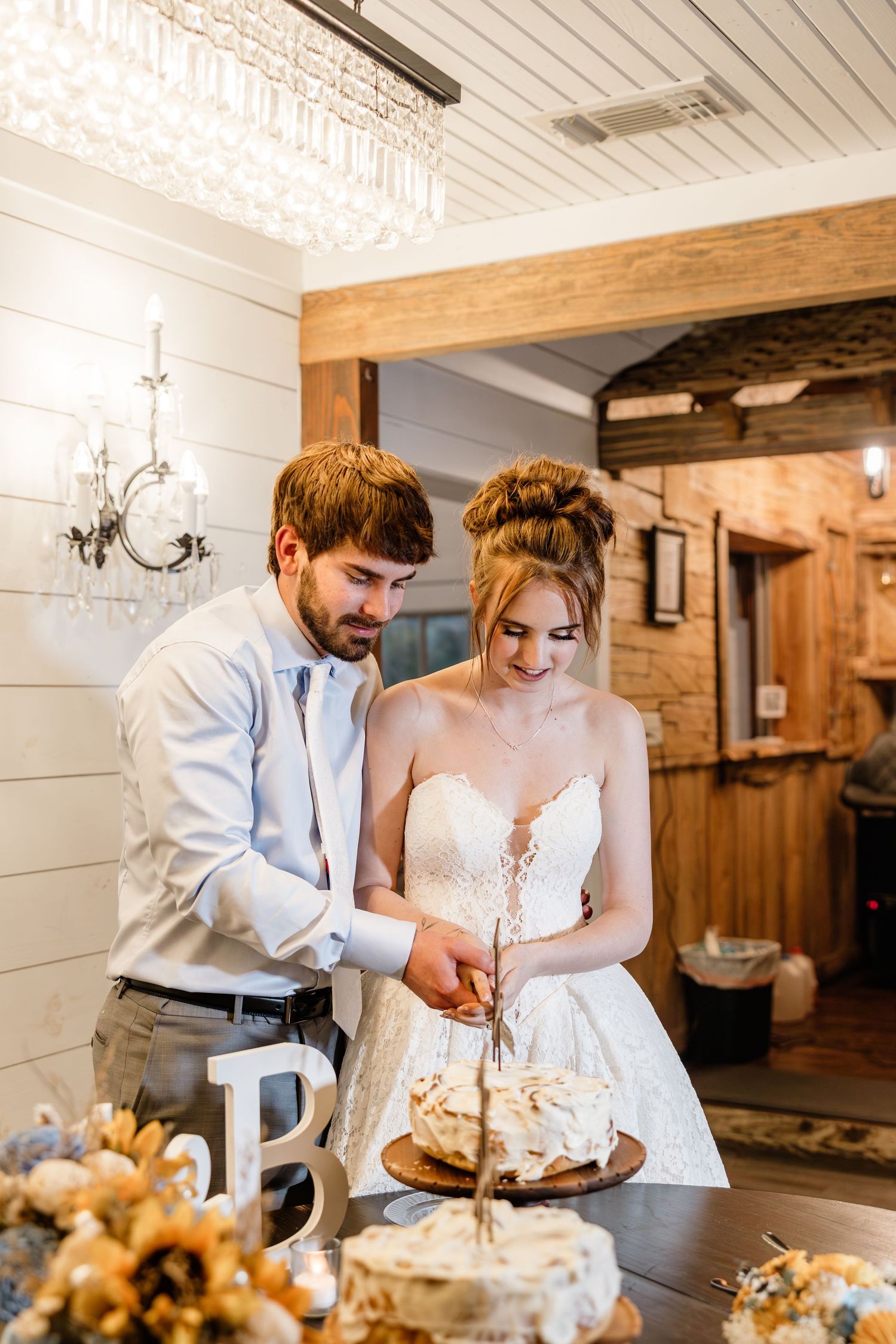 Bride and groom cutting their rustic wedding cake together in a charming barn venue with elegant chandelier lighting.