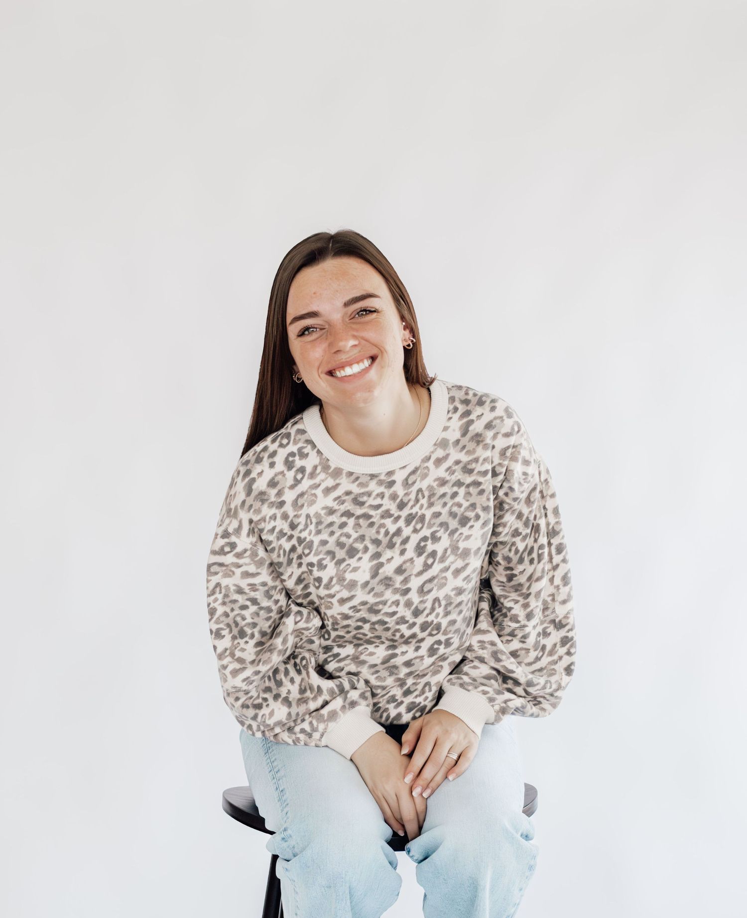 A person wearing a cozy leopard print sweater and light blue jeans sits and smiles against a white background.