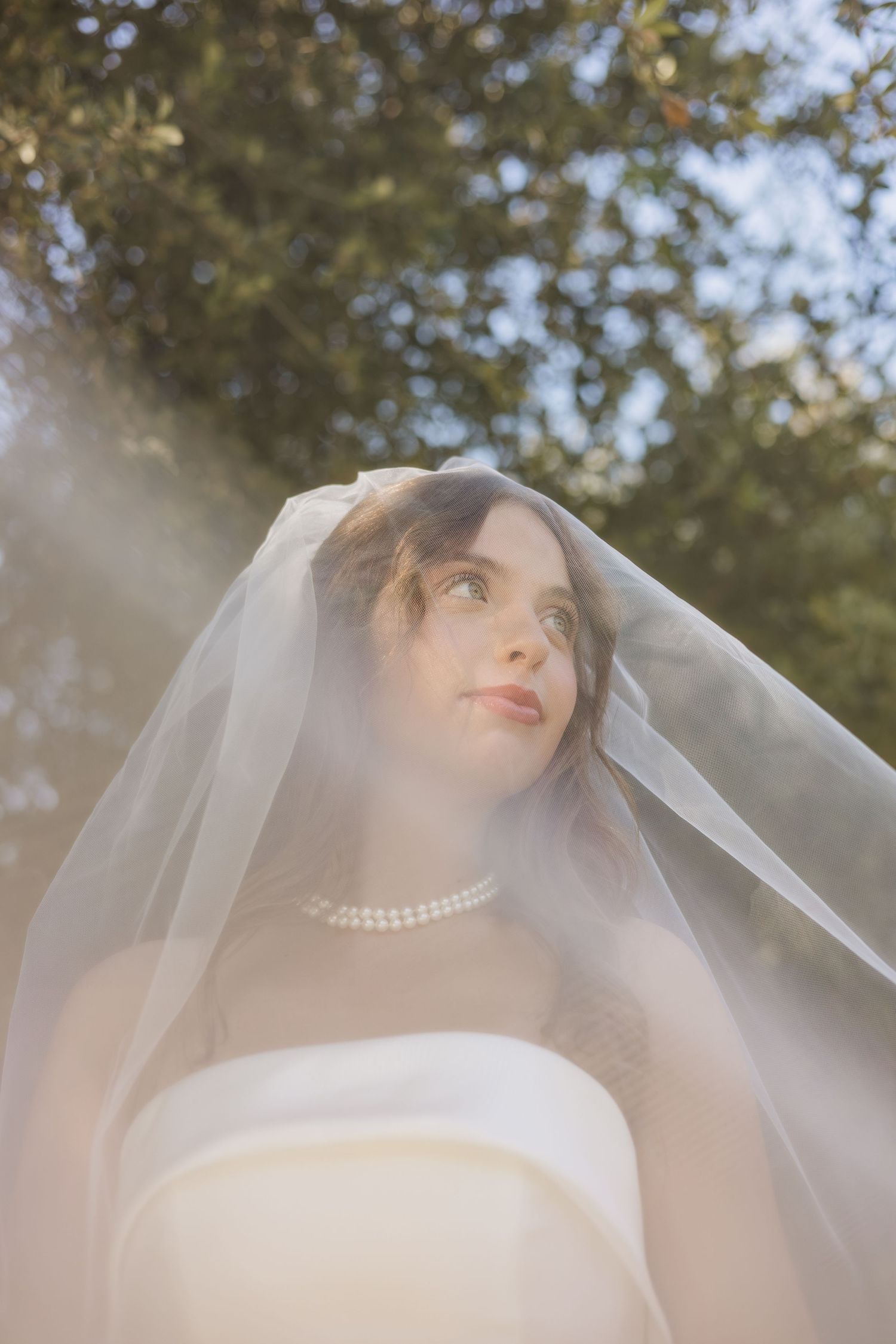 Bride in white gown and veil gazes upward outdoors, sunlight filtering through trees, wearing pearl necklace.