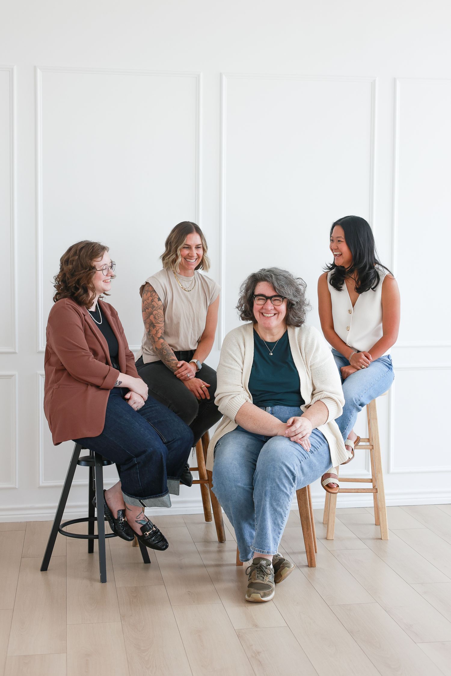 Four women laughing and sitting on stools against a white wall in a bright, modern studio setting.
