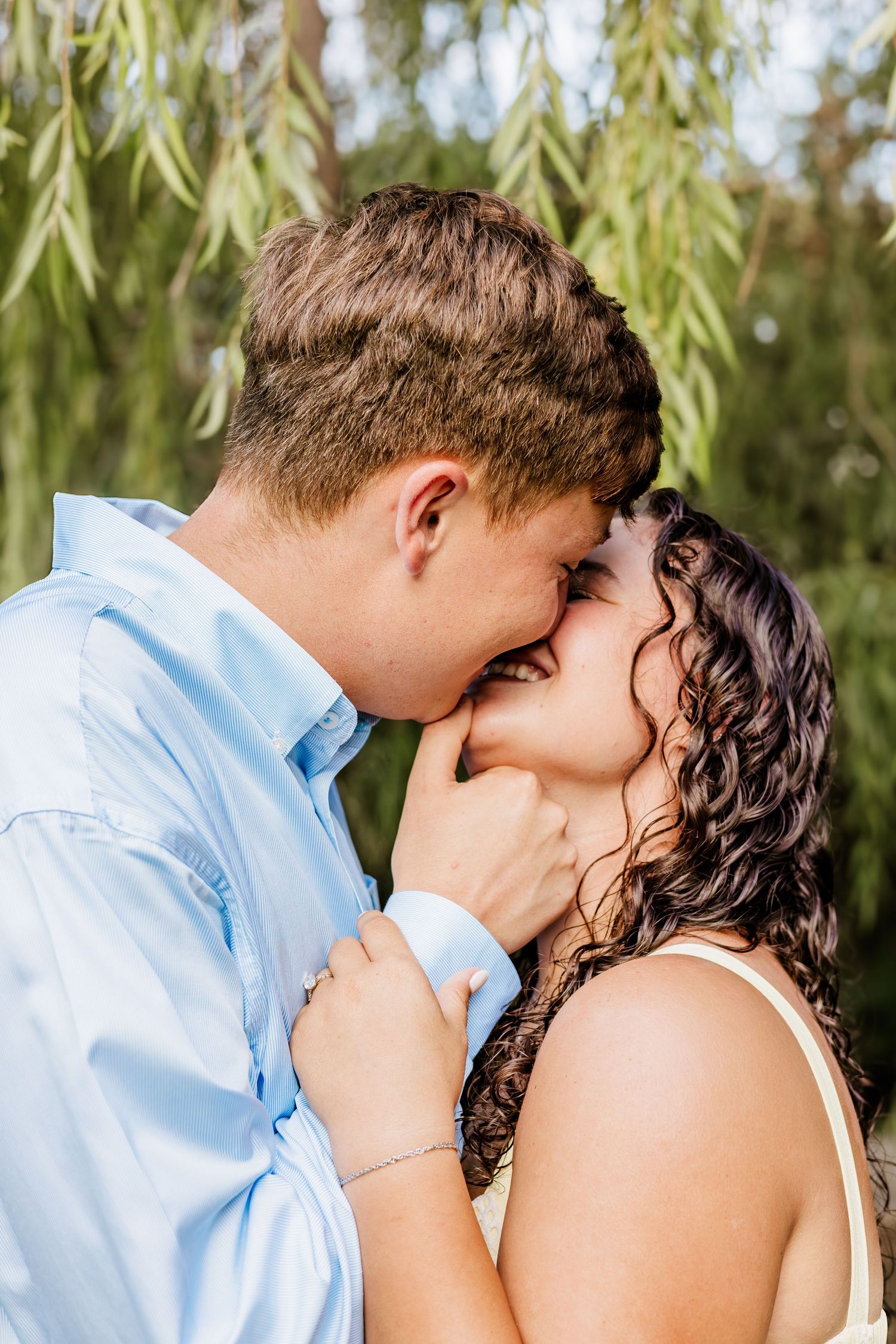 A couple sharing a romantic kiss under weeping willow branches outdoors.