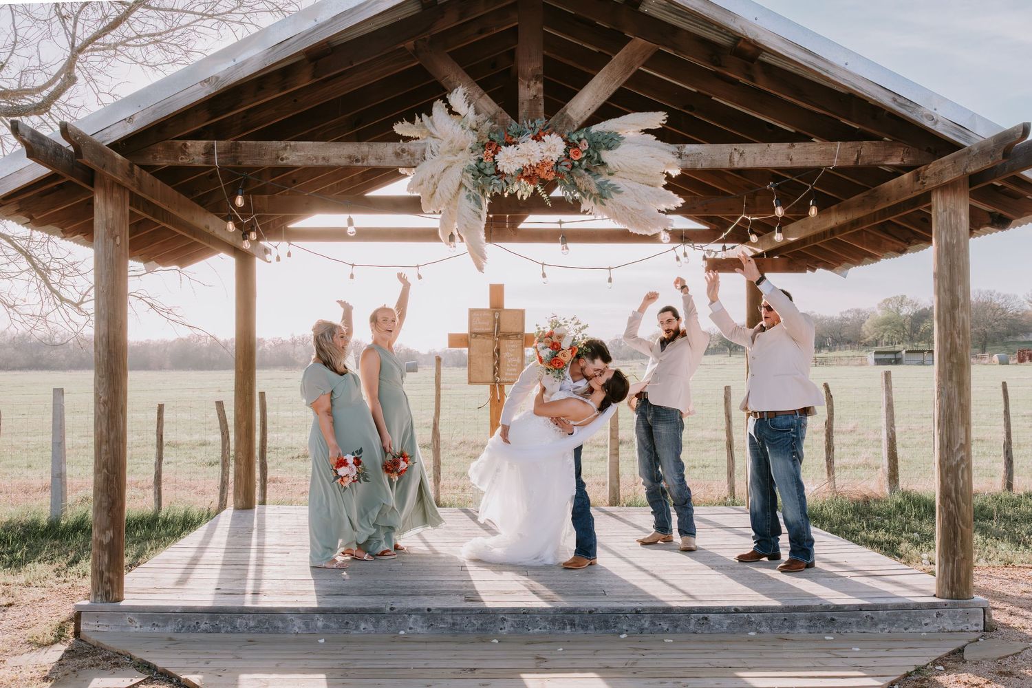 Wedding party celebrates under a rustic wooden gazebo on a farm, with bride and groom kissing surrounded by bridesmaids and groomsmen.