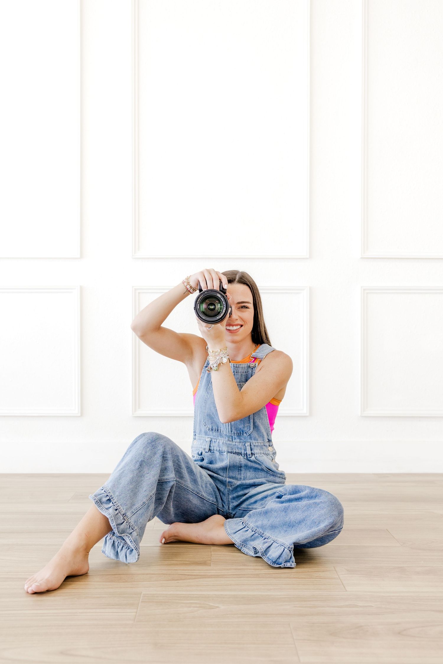 Young woman in denim overalls sitting on floor holding camera up to shoot, smiling in bright white room.