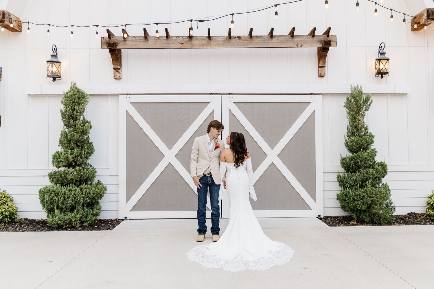 Bride and groom share a romantic moment in front of a white barn venue with string lights and topiaries.
