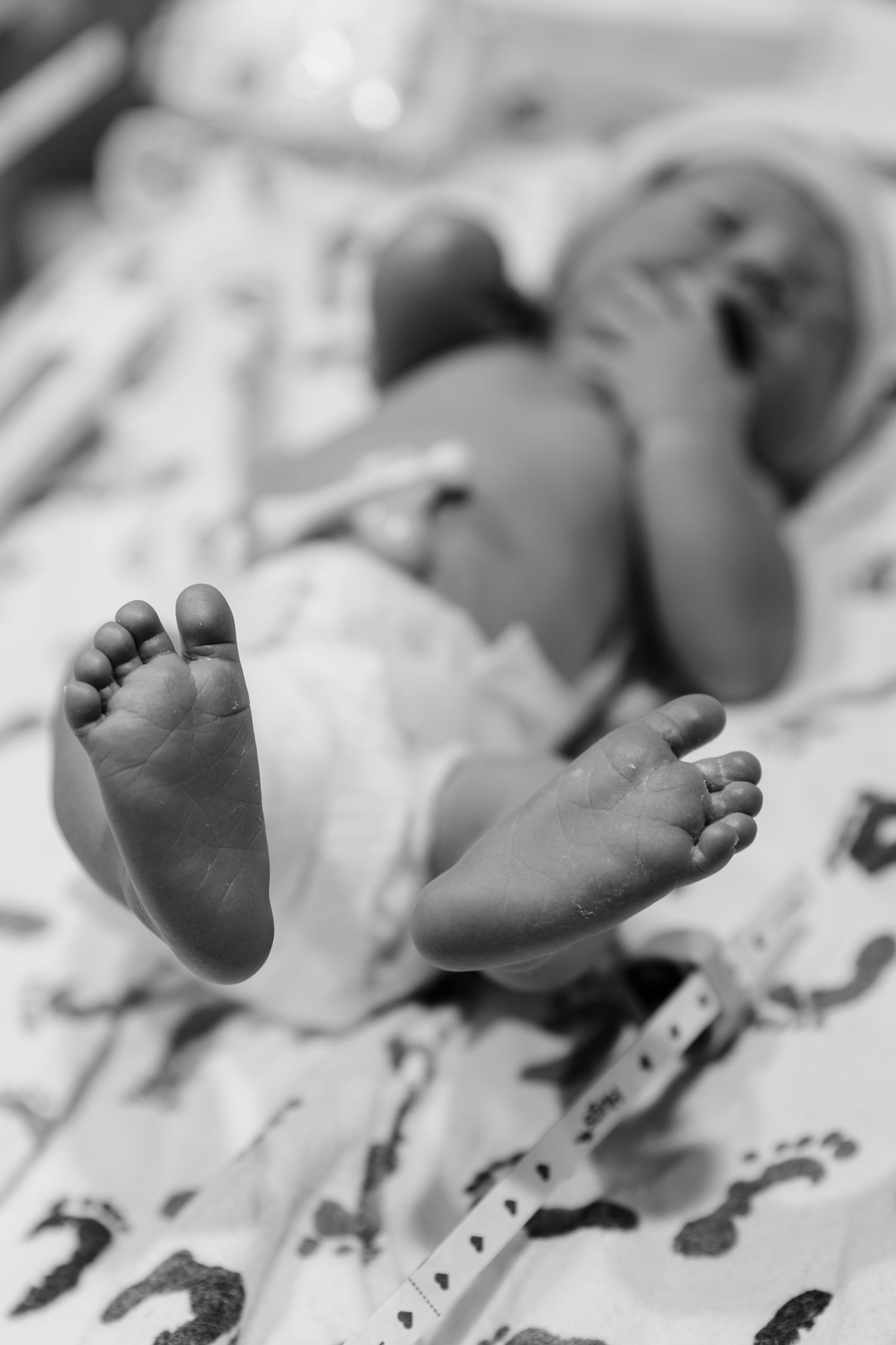 Black and white close-up photo of a newborn baby's tiny feet lying on a patterned hospital blanket.